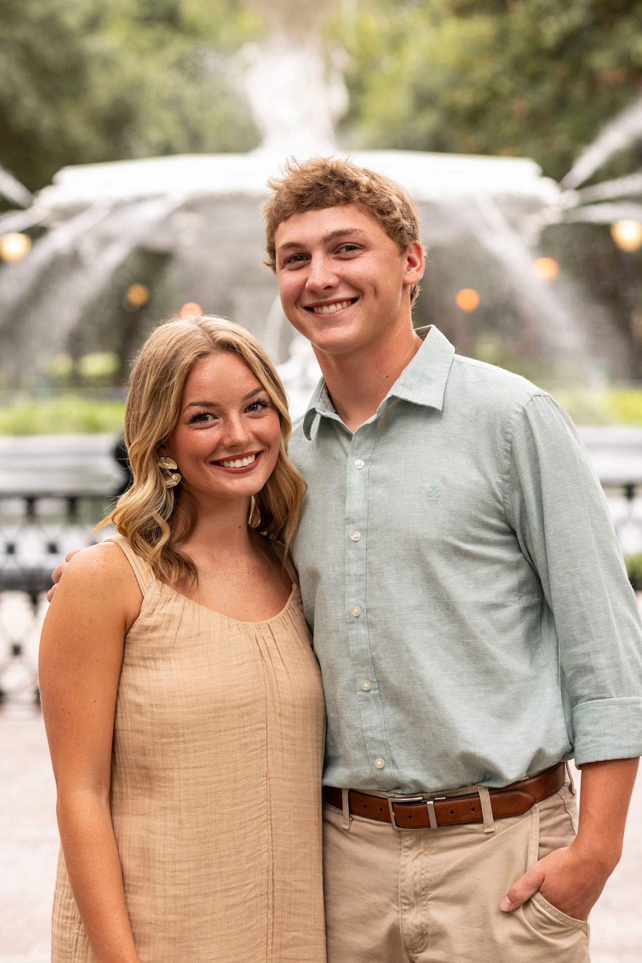 A couple poses outdoors by a fountain wearing summer attire with khakis and beige dress.