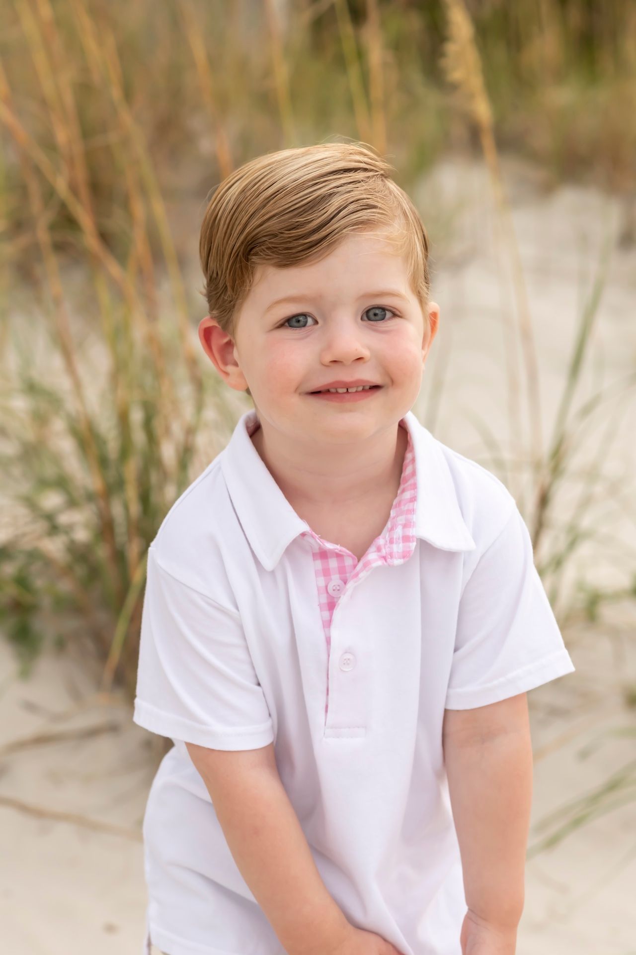 Child wearing white polo shirt sits smiling on sandy beach with beach grass in background.