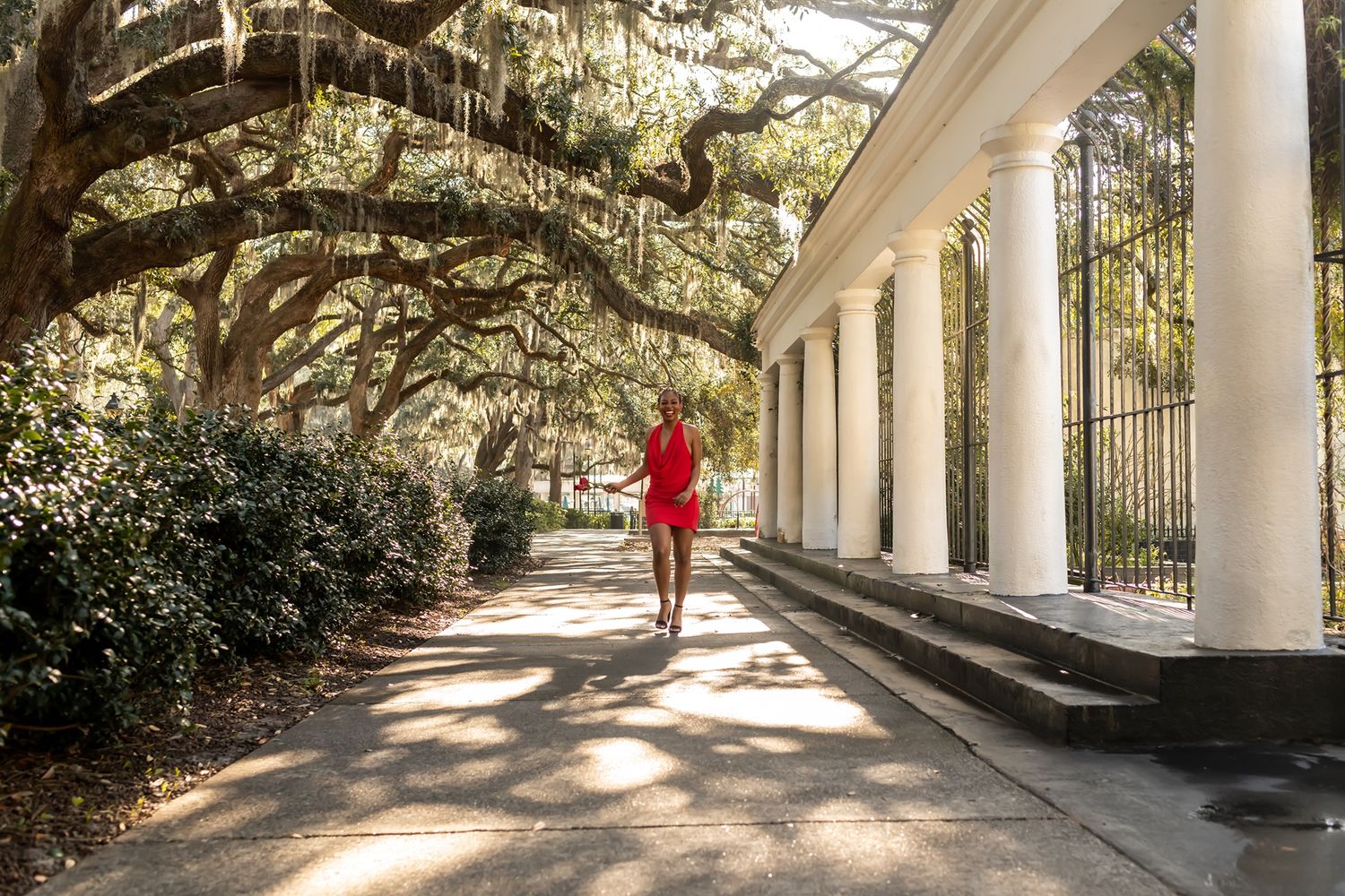 A person in a red dress walks along a covered walkway with white columns and overhanging Spanish moss at a Southern estate.