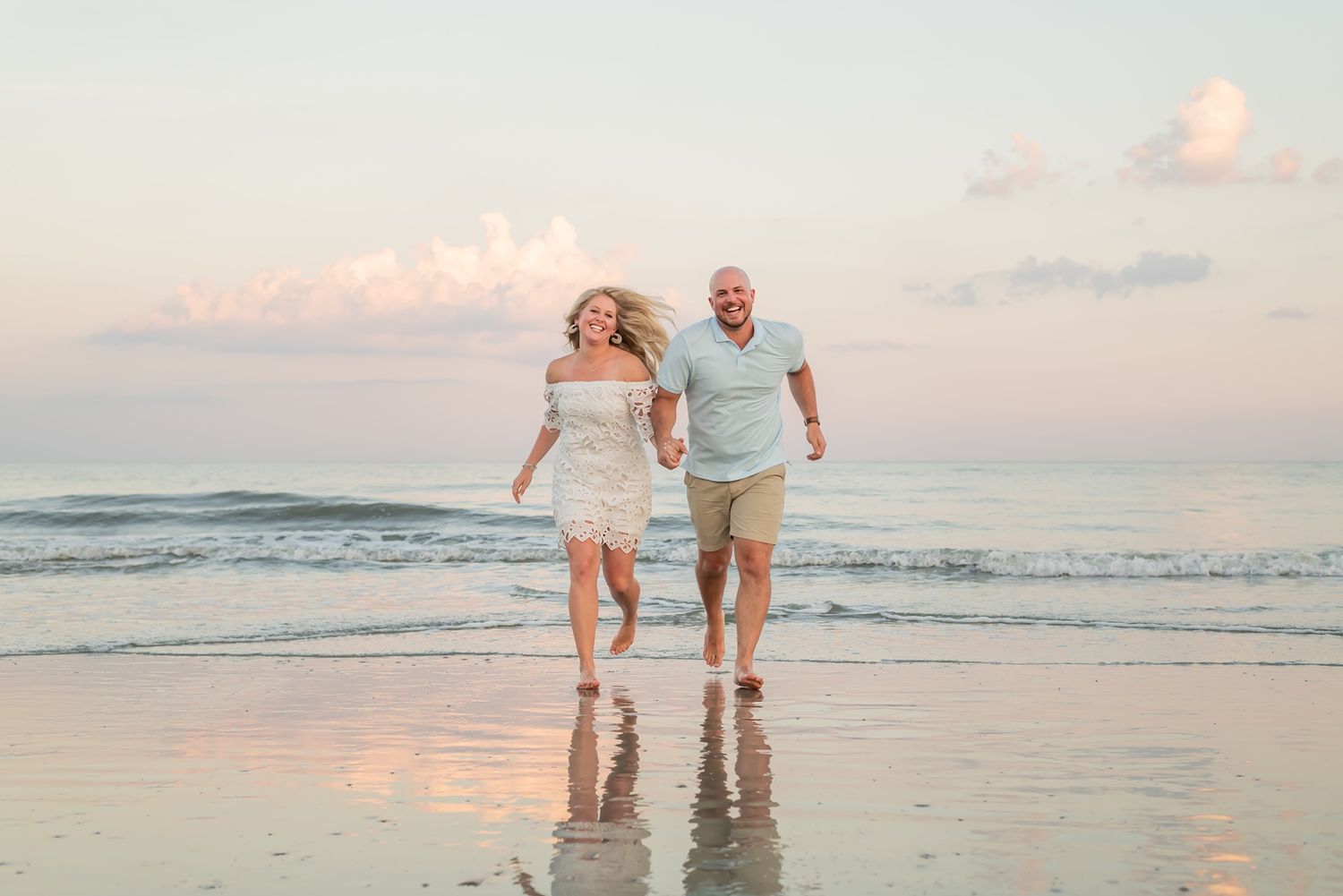 A couple runs joyfully along the beach at sunset, splashing through shallow water with soft pink skies in the background.