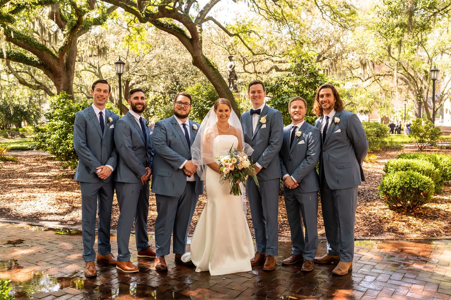Wedding party in gray suits poses on brick pathway under oak trees in a garden setting.