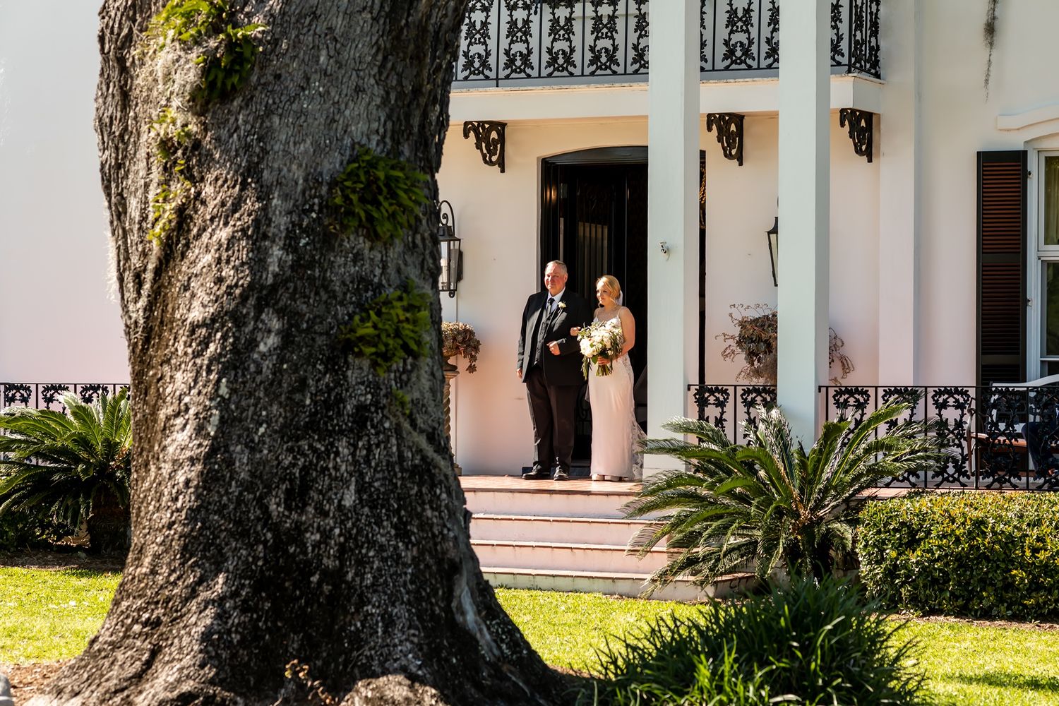 Historic white building facade with wrap-around porch, tall windows, and lush landscaping in tropical setting.