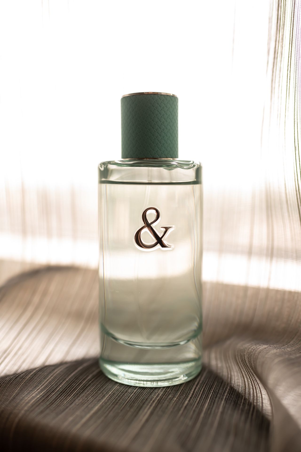 Clear glass perfume bottle with an ampersand symbol and green cap sitting on a textured surface in soft lighting.