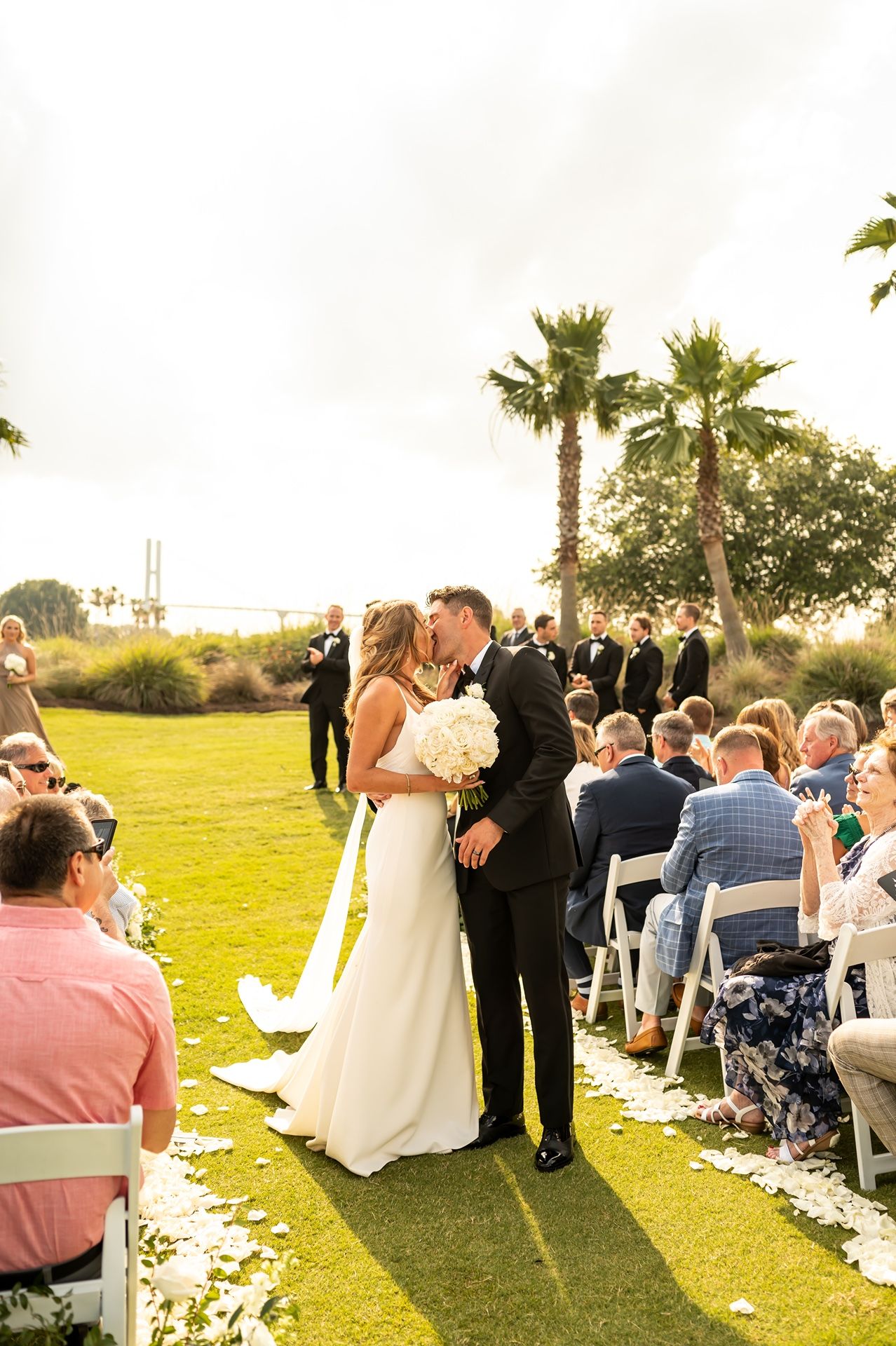 Couple sharing first kiss during outdoor wedding ceremony on grassy lawn with palm trees and guests looking on.