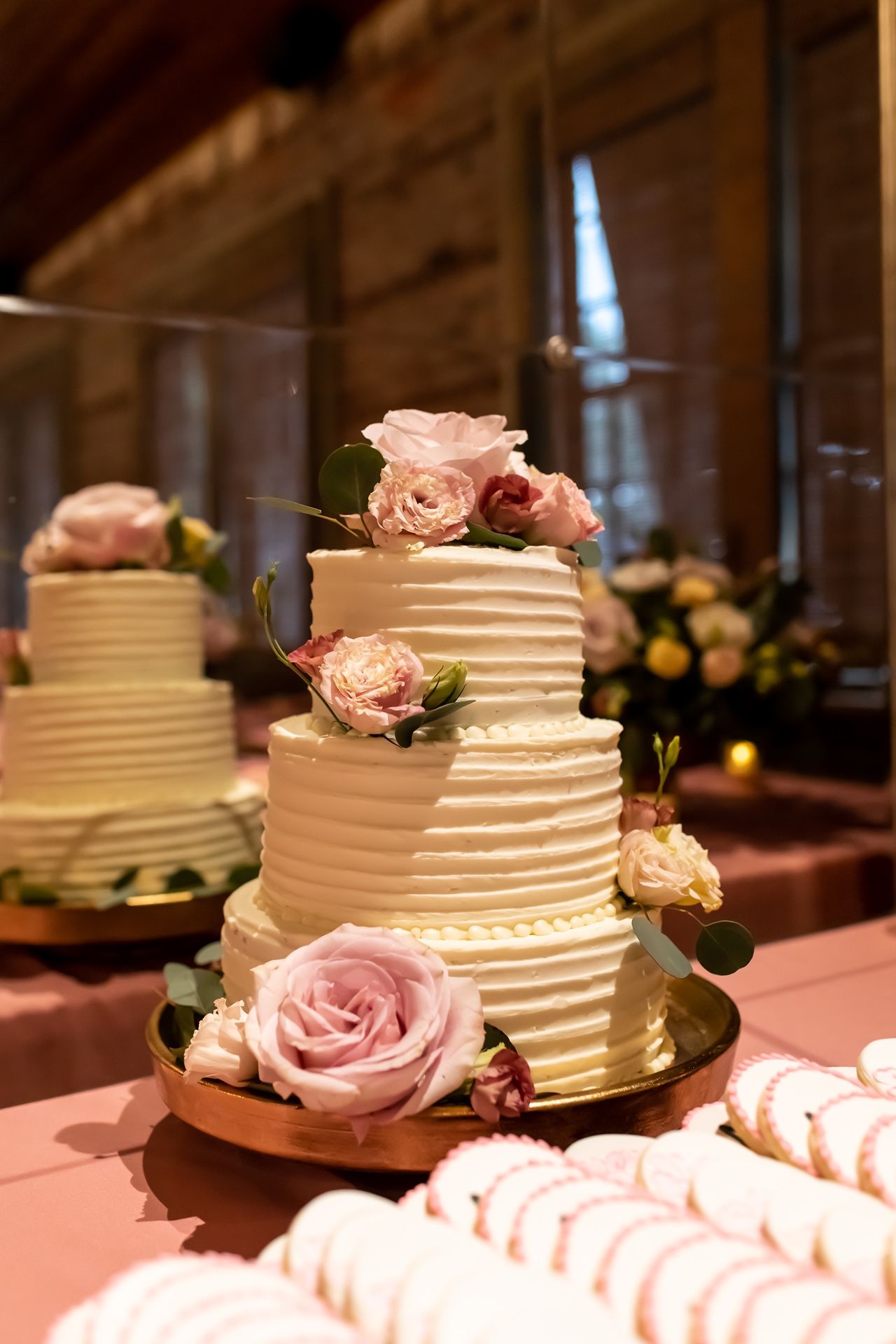 Elegant three-tier white wedding cake decorated with soft pink roses and textured buttercream stripes on a peach tablecloth.