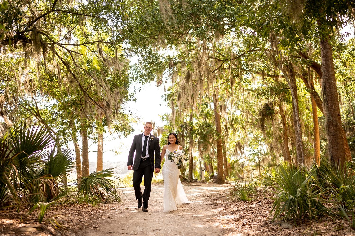 A couple walks hand in hand along a scenic path beneath Spanish moss-draped oak trees in a natural forest setting.