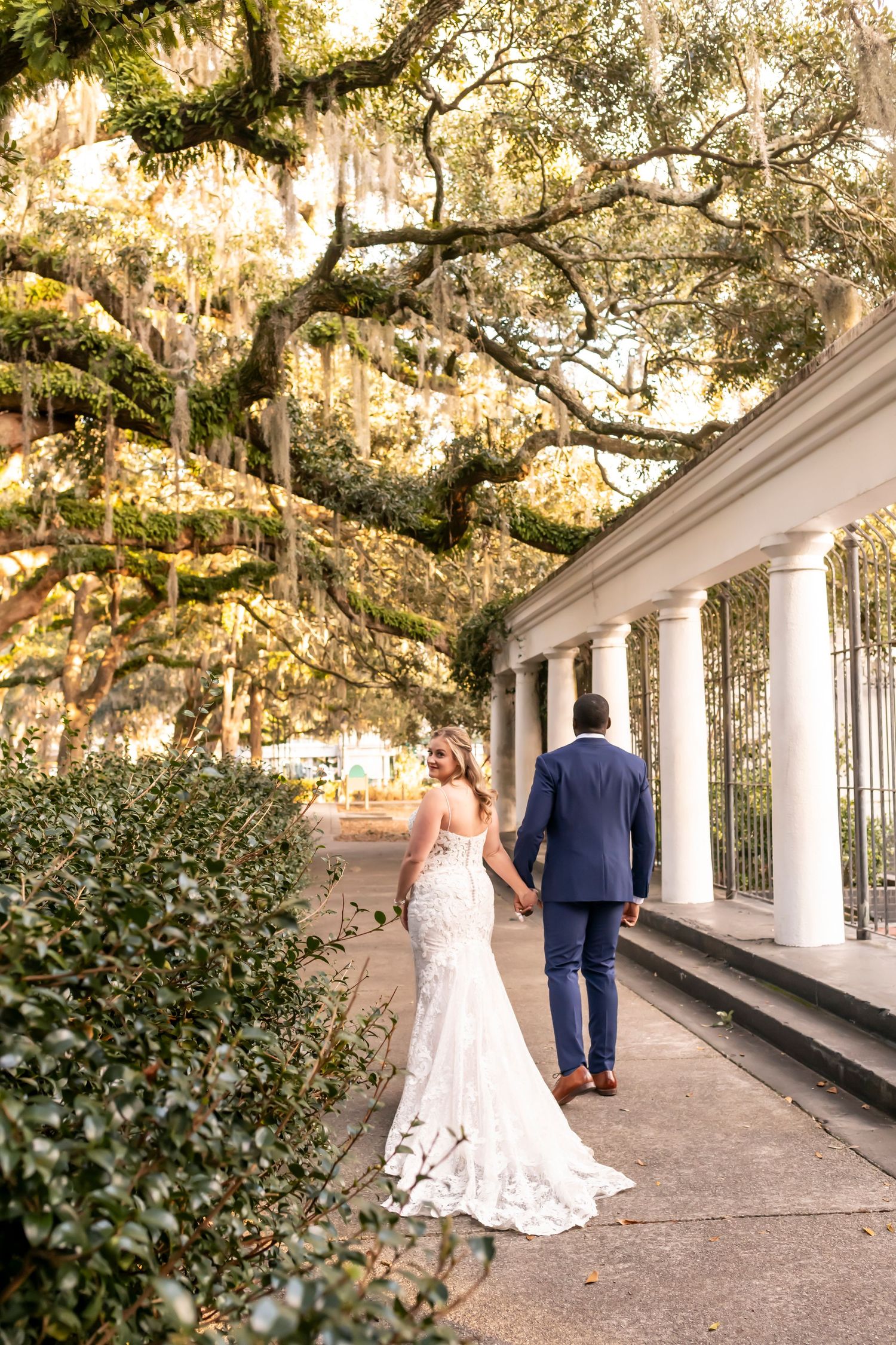 A couple strolls along a white columned walkway beneath beautiful Spanish moss-draped oak trees in a romantic garden setting.