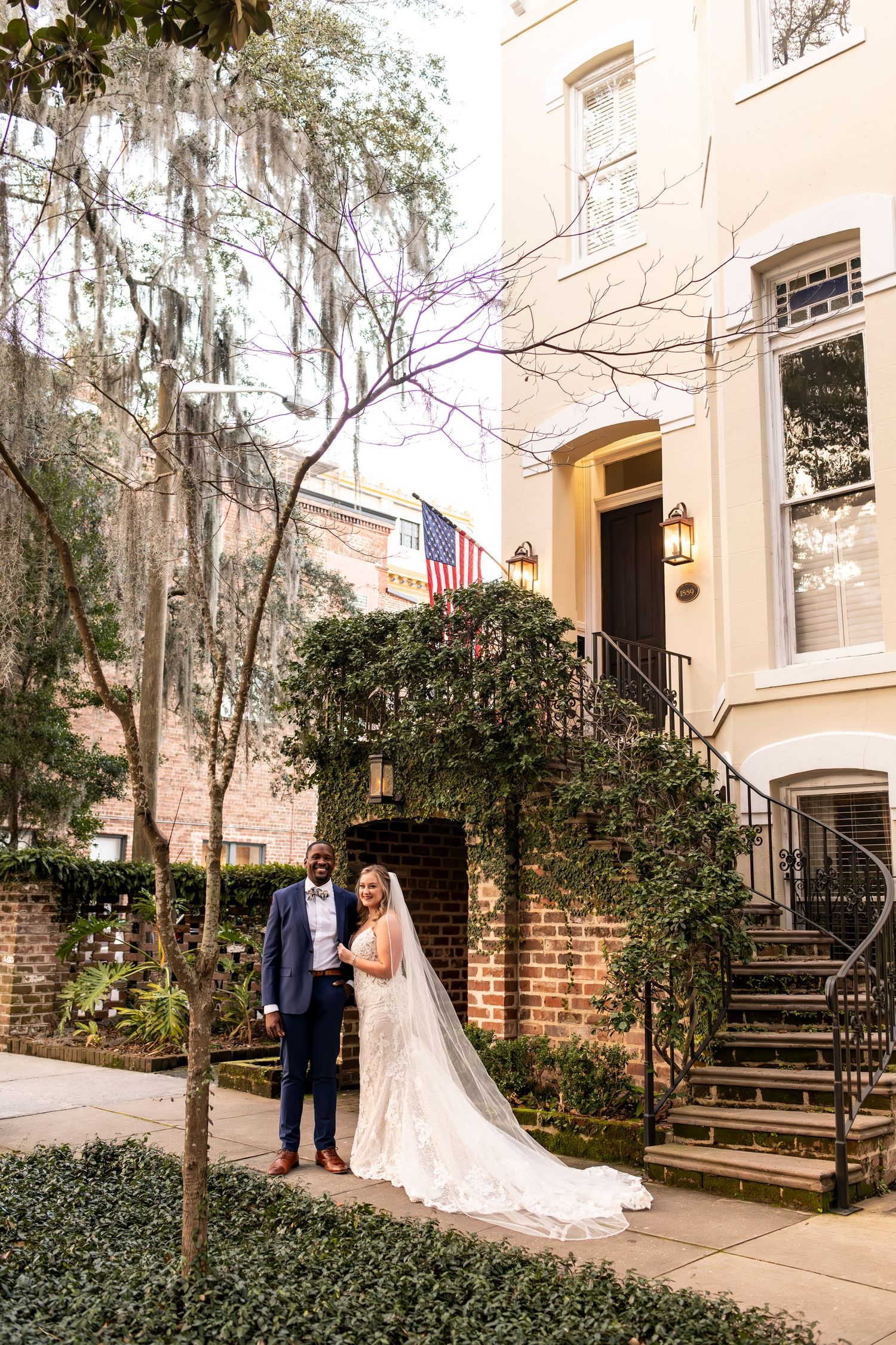 A couple stands in front of a historic Southern building with Spanish moss and American flag in Savannah, Georgia.