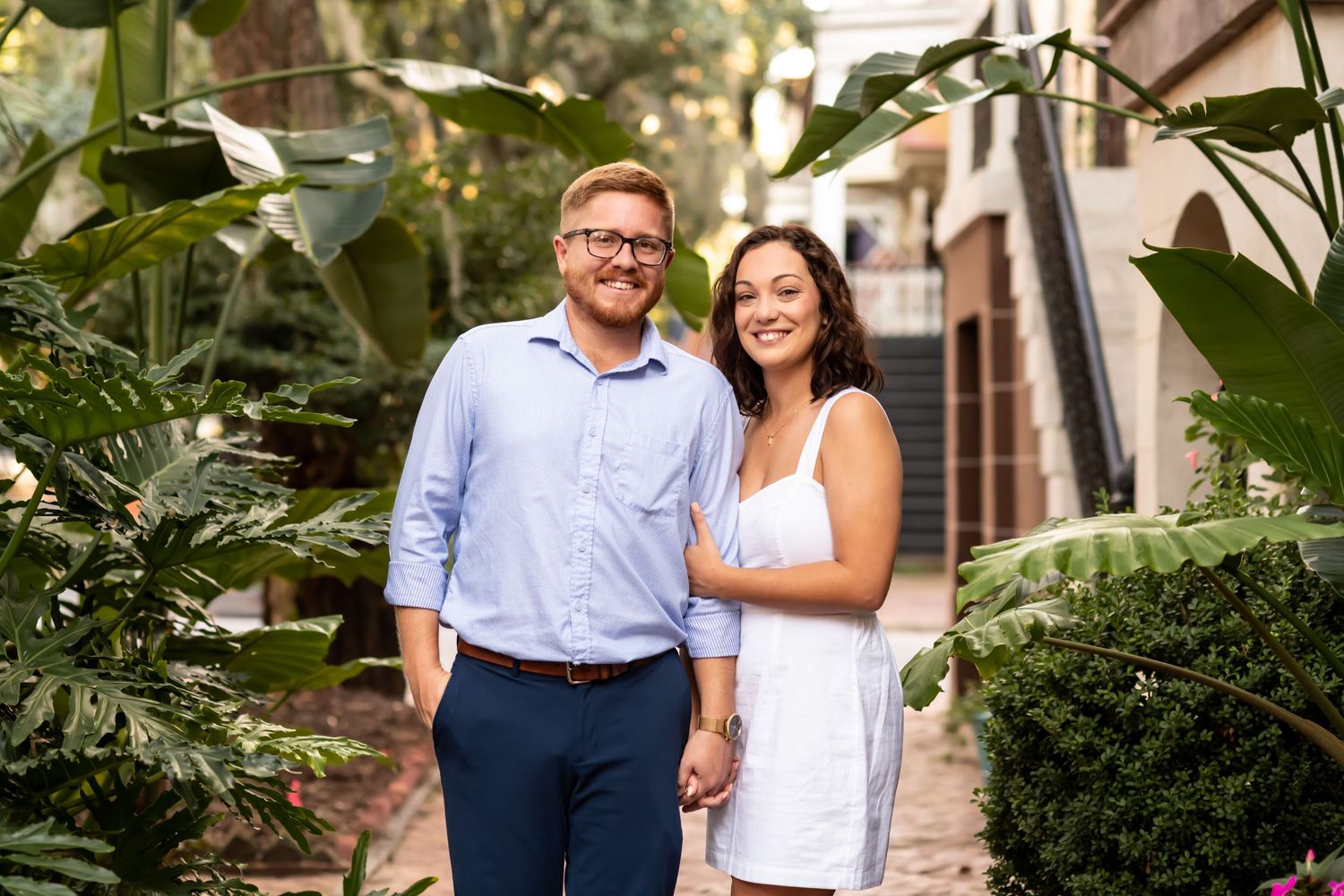 A couple in semi-formal attire stand together on a lush garden path lined with tropical plants and brick walls.