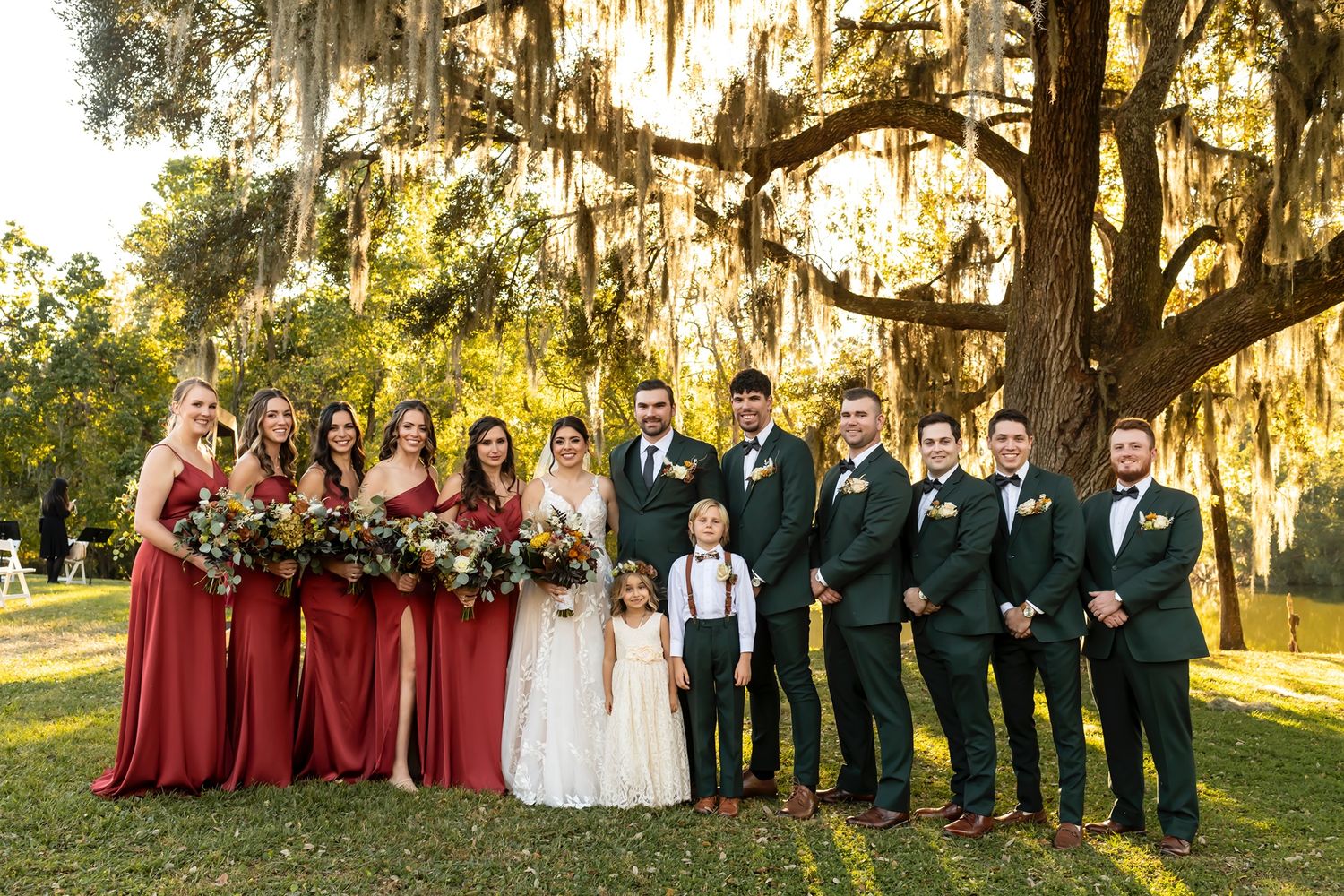 Wedding party poses outdoors under Spanish moss draped oak trees at golden hour in garden setting.
