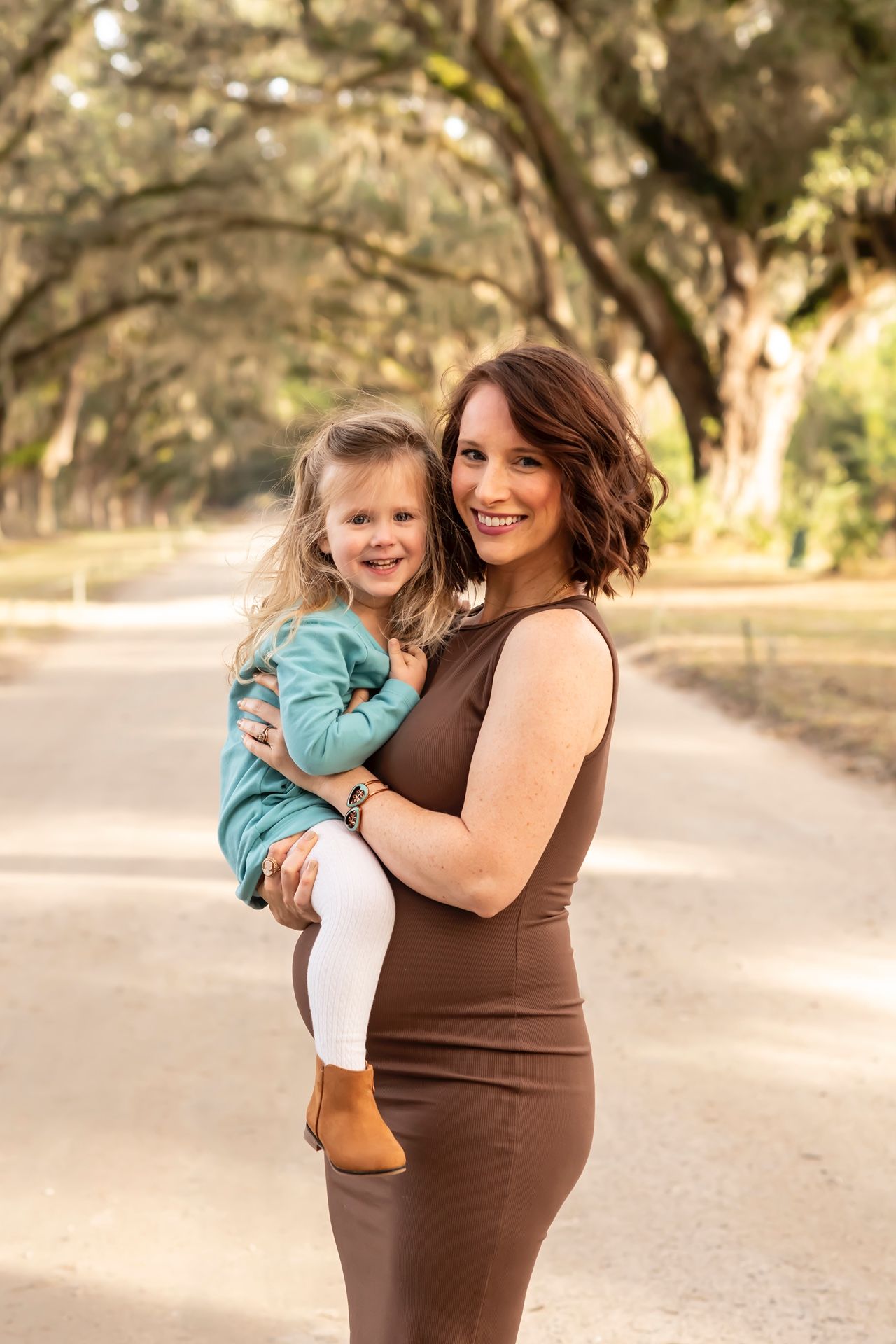 Mother and child in brown dress share playful outdoor moments during sunset in a tree-lined path.