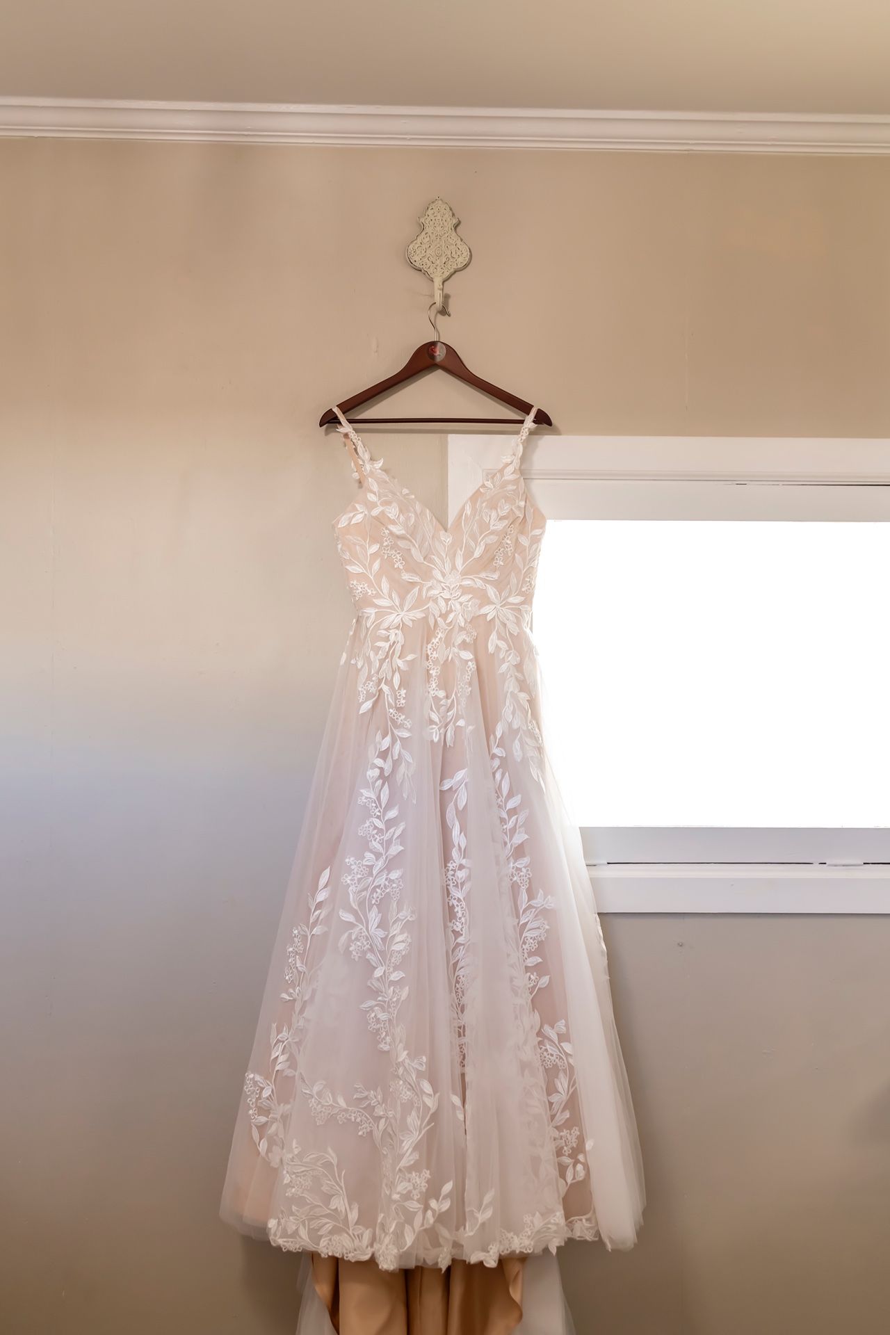 A delicate lace wedding dress with floral appliques hangs from a decorative hanger against a beige wall near a window.