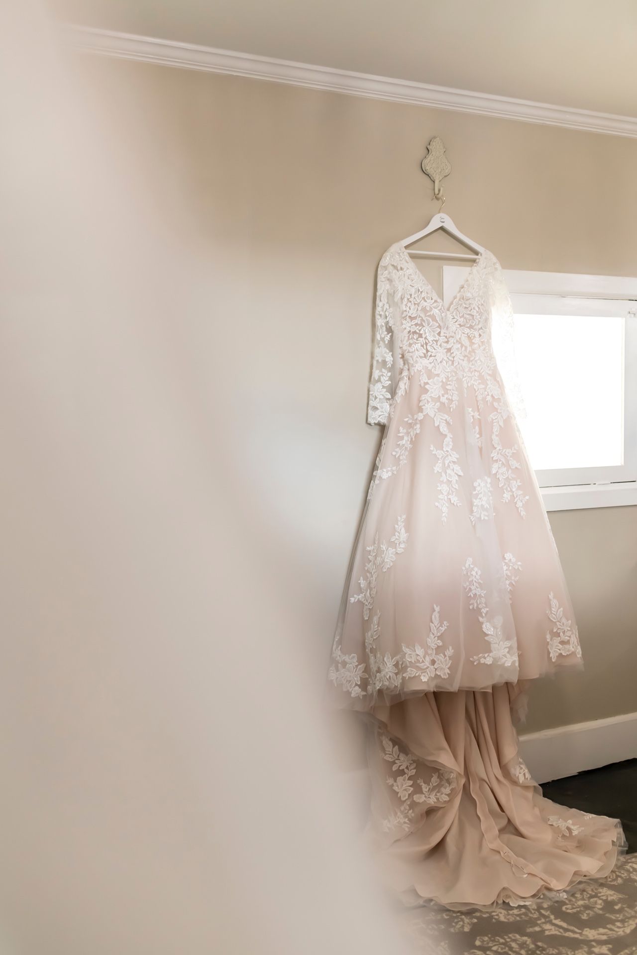 A delicate white lace wedding dress hangs from a hanger against a cream-colored wall near a window.