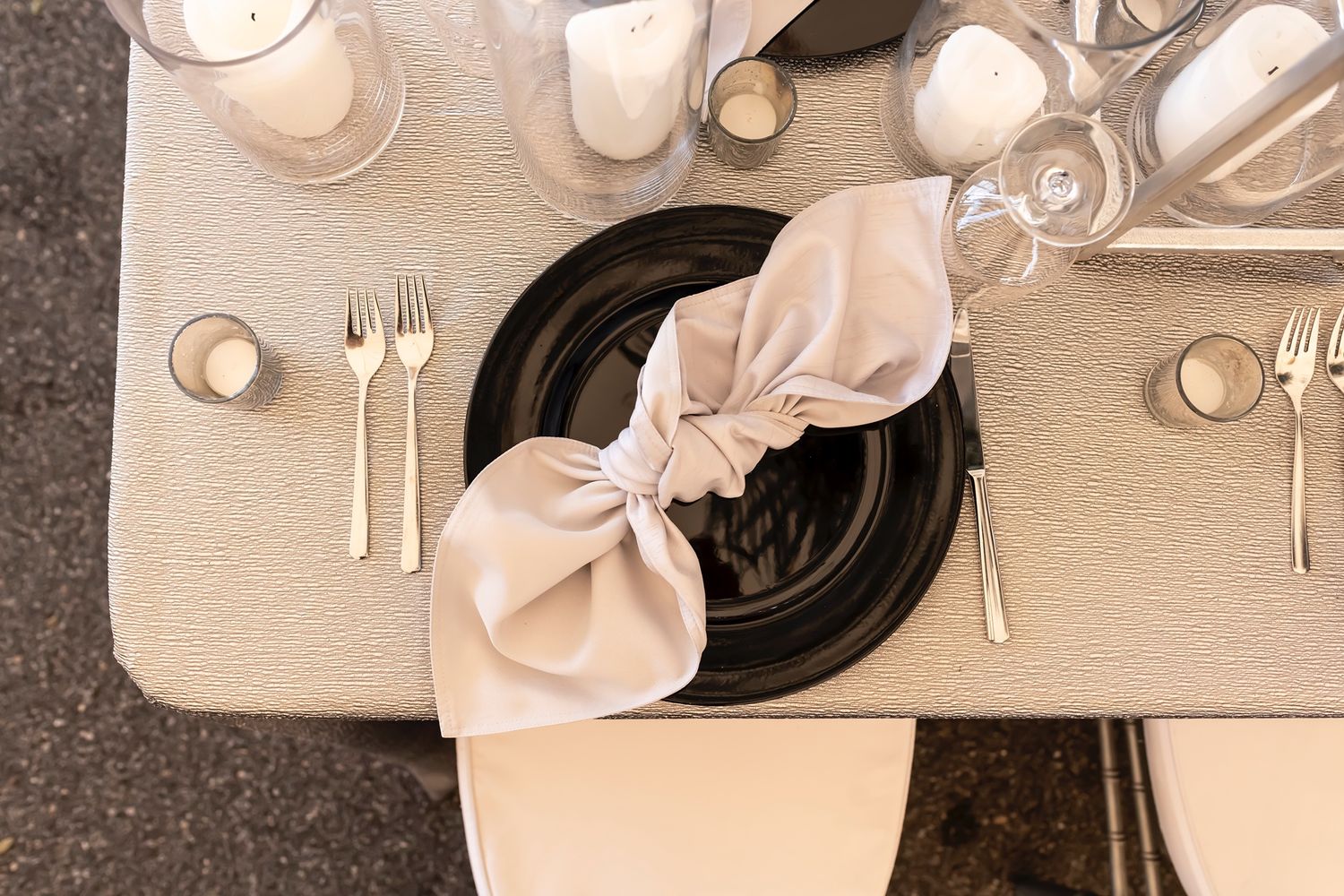 Elegant table setting featuring a black plate with beige bow napkin, silverware and glasses on textured cream tablecloth.