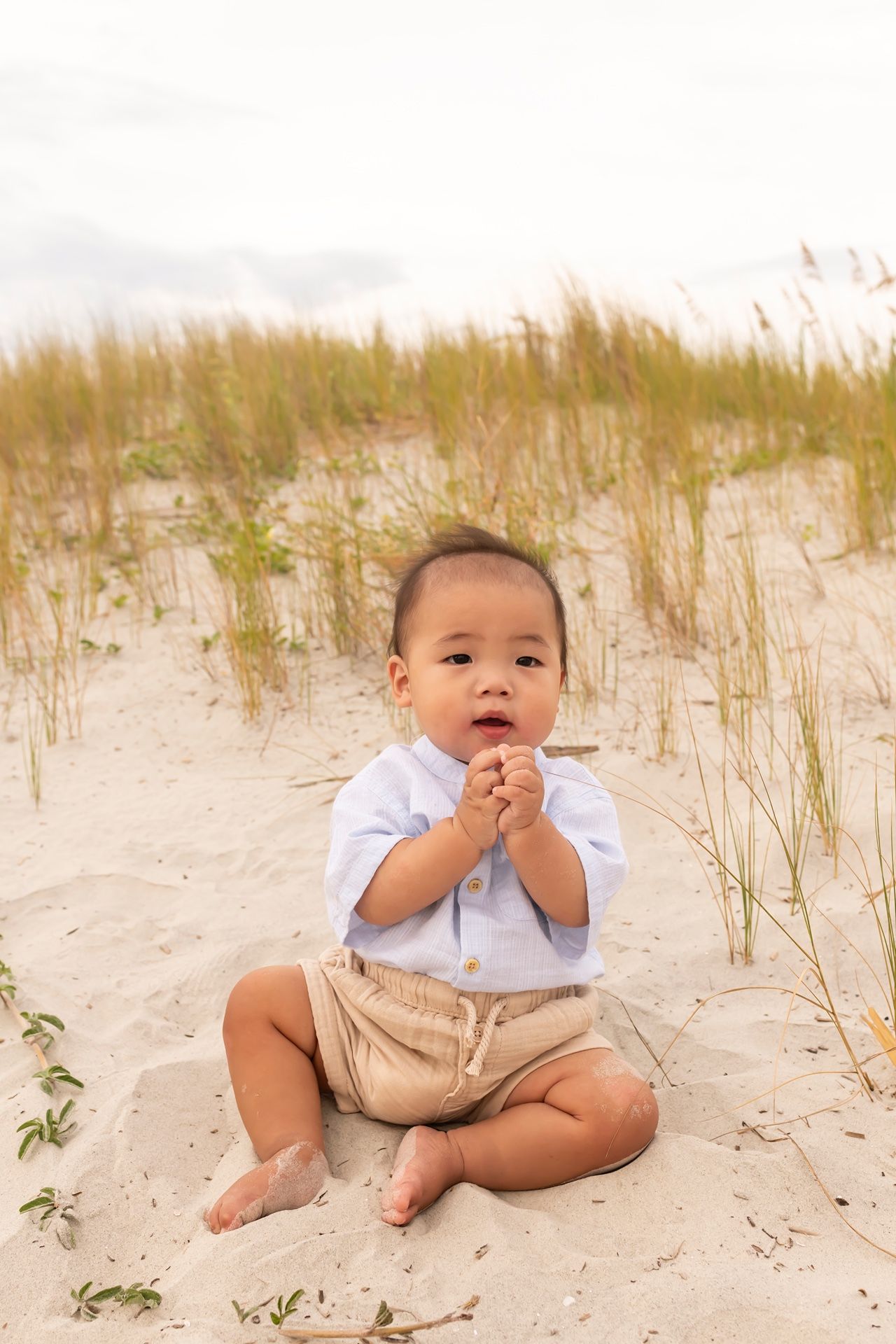 Small child in white shirt and khaki shorts sitting on sandy beach dunes with grass in background.