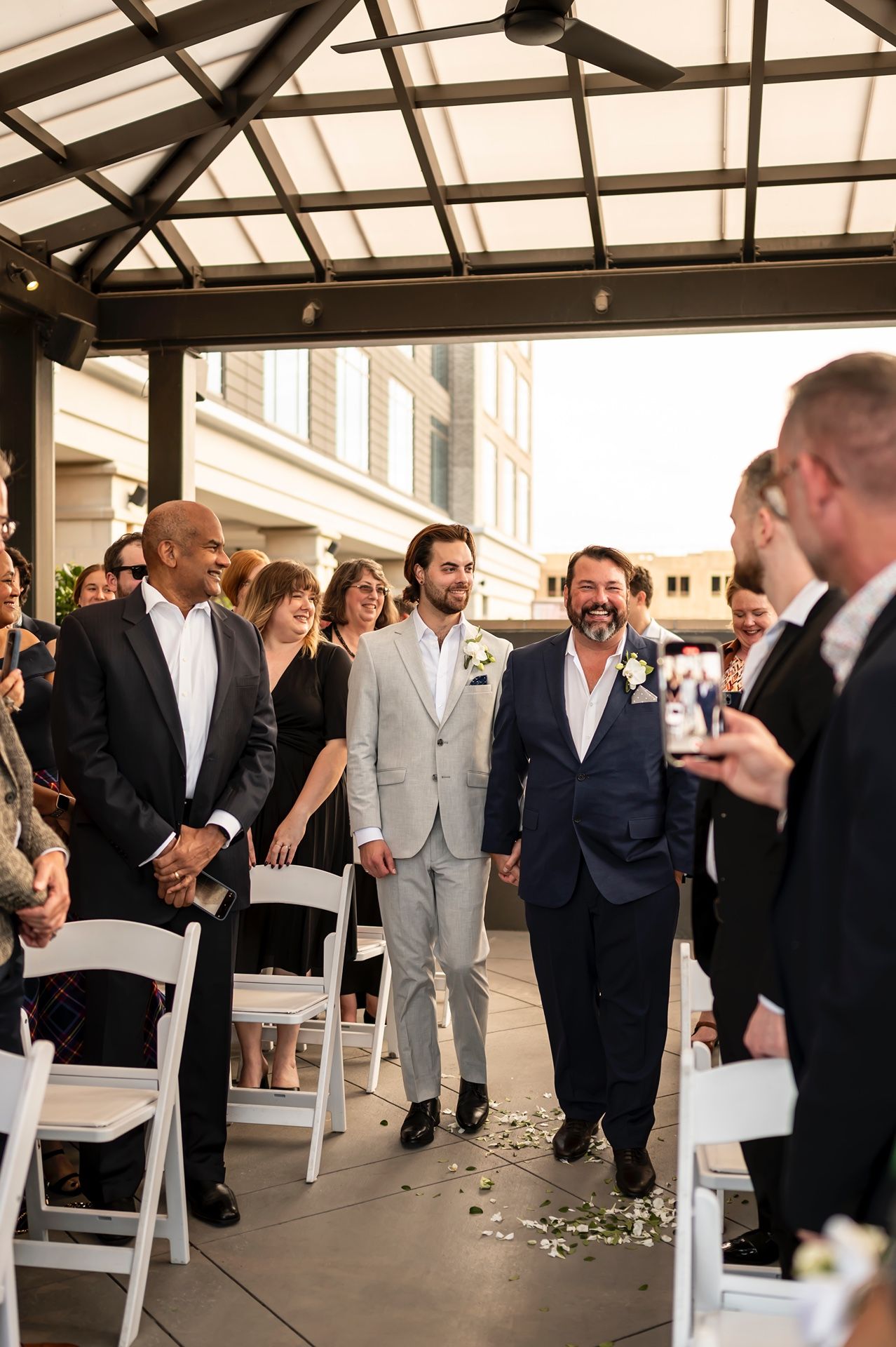 Wedding guests celebrate on a covered terrace with white chairs and scattered flower petals during a reception.