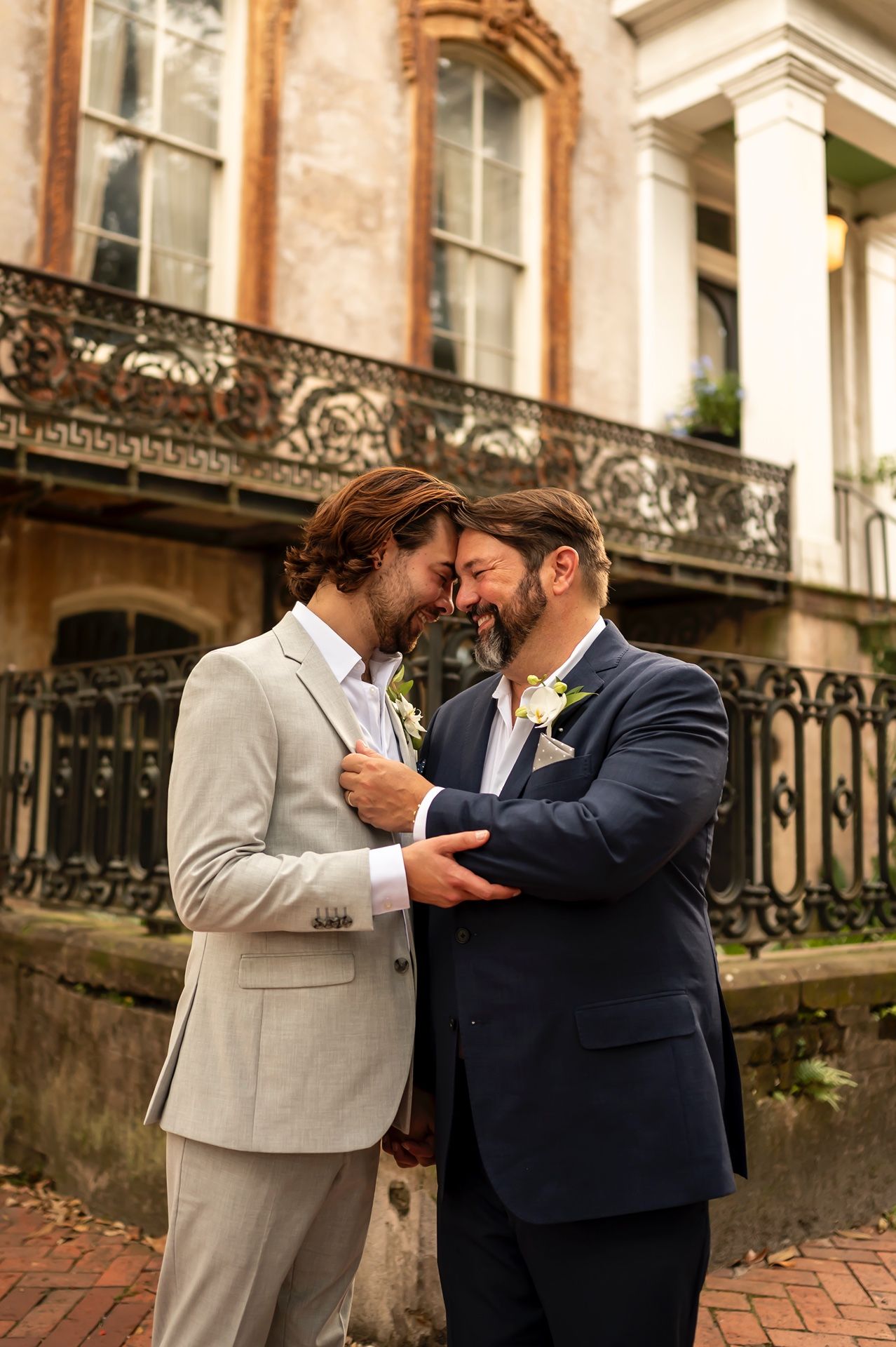 Two grooms sharing an intimate moment in front of a historic building with ornate ironwork details.