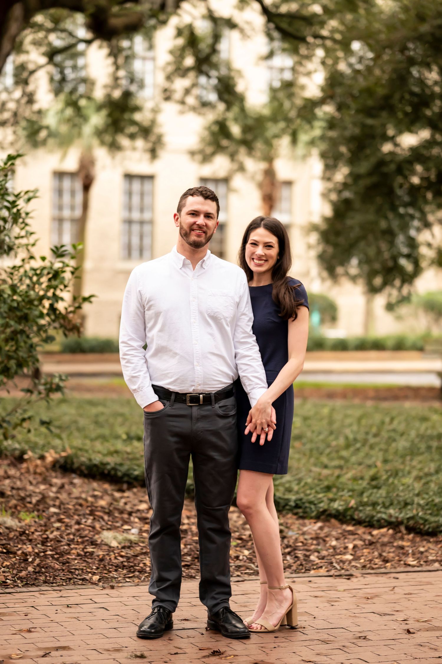 A couple stands together on a brick pathway with a building in the background during an outdoor portrait session.