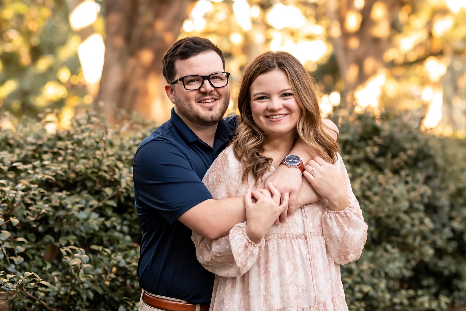 A couple poses together outdoors during golden hour with lush greenery and warm sunlight in the background.