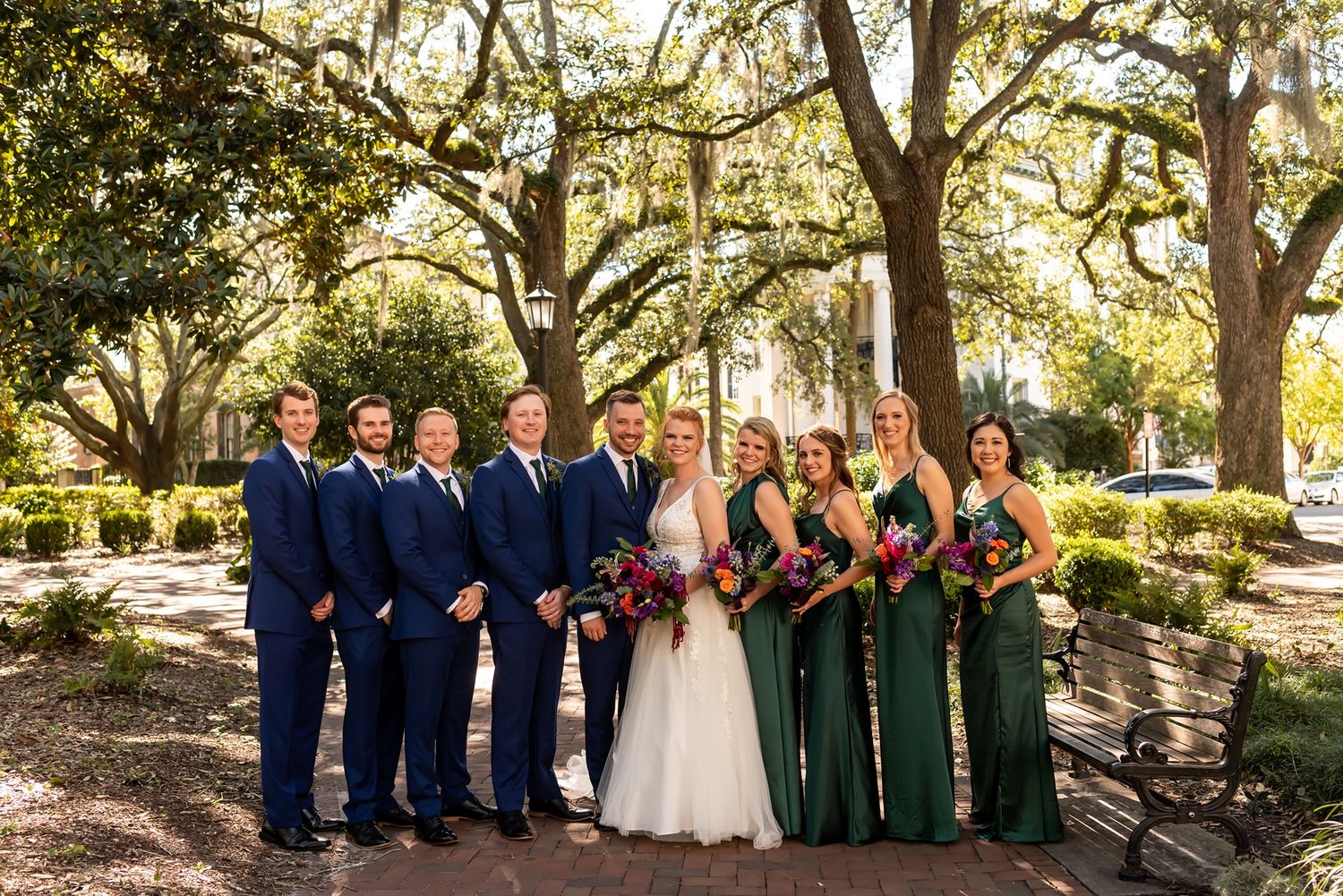 Wedding party poses under oak trees in southern garden setting with bridesmaids in green and groomsmen in navy suits.