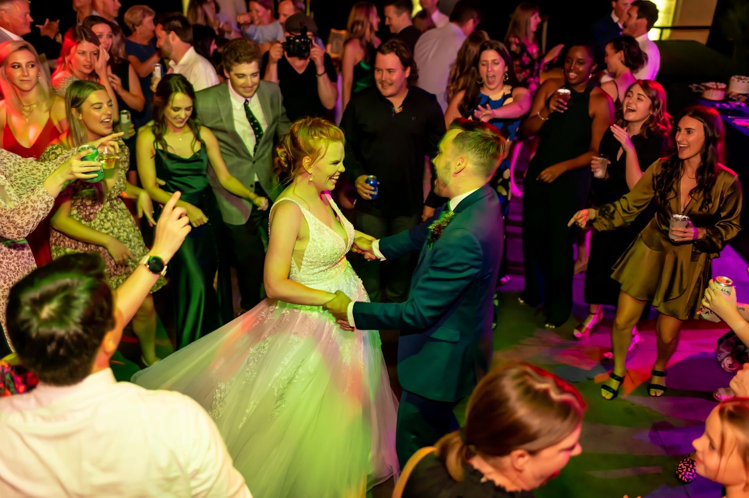 Wedding guests surround a couple dancing at a reception with colorful lighting creating a festive party atmosphere.