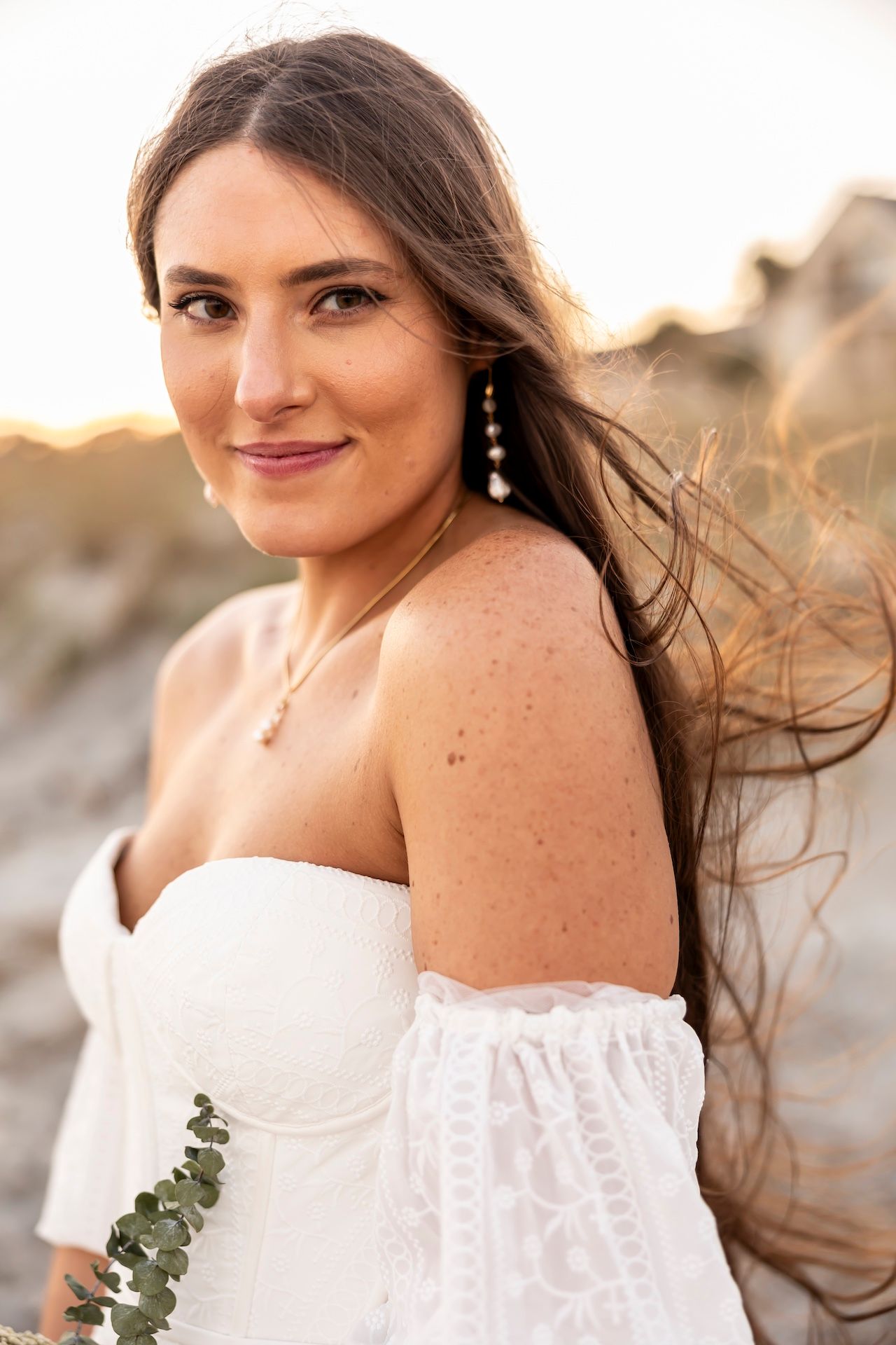 Woman in white off-shoulder wedding dress with long flowing hair poses at sunset on a beach.