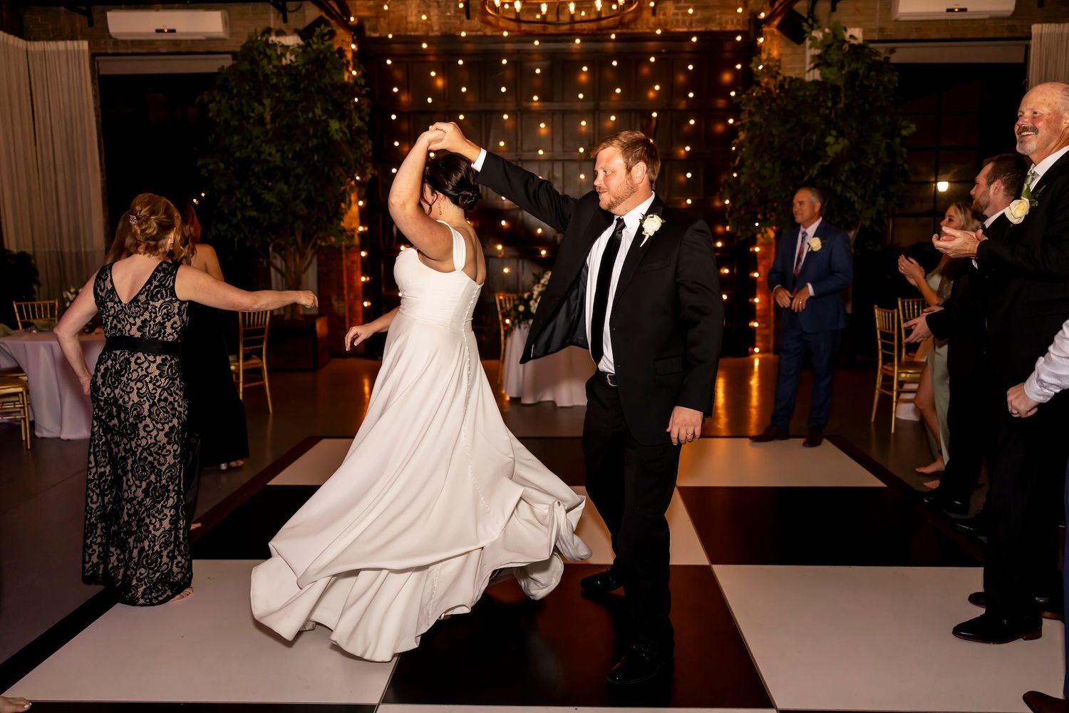 A couple dances together on a black and white checkered dance floor at an elegant reception venue with twinkle lights.