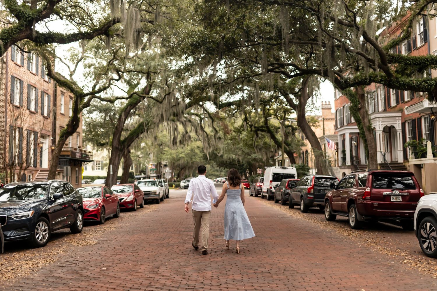 A tree-lined brick street in a historic neighborhood with parked cars and two people walking together.