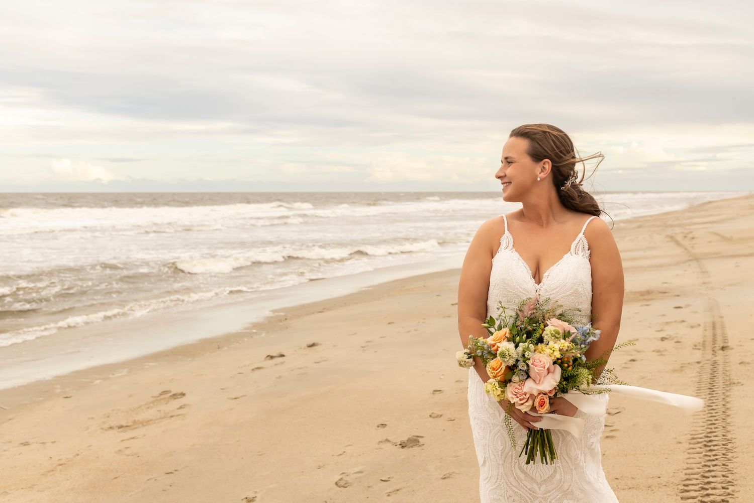 Someone in a white wedding dress holds a colorful bouquet while standing on a sandy beach at sunset.