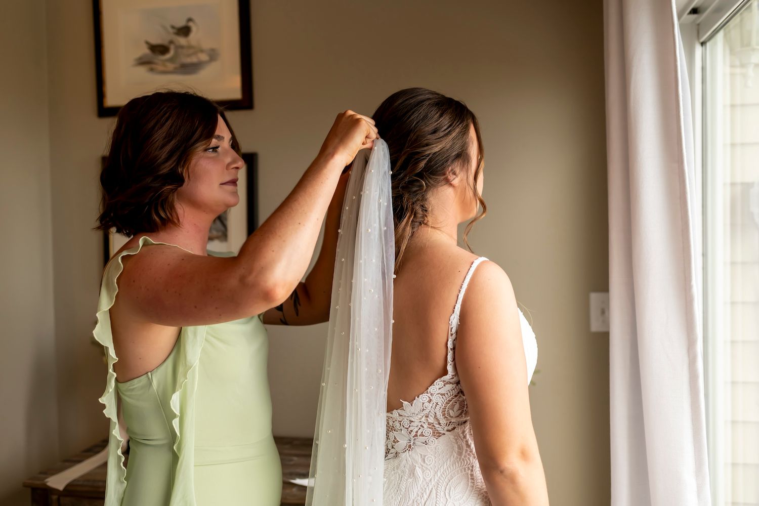 A helper adjusts a long white wedding veil near a bright window in a getting-ready room.