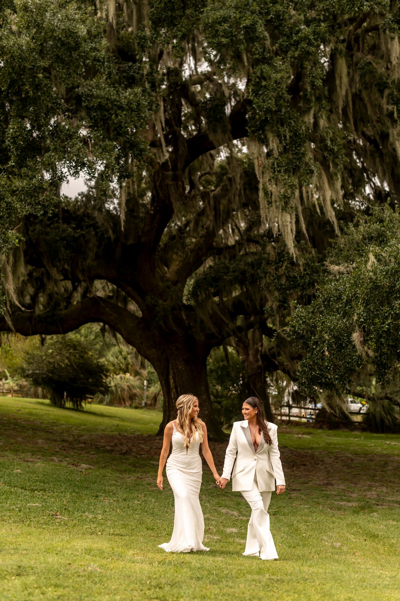 Two people in white wedding attire walk together under majestic Spanish moss-draped oak trees on a grassy lawn.