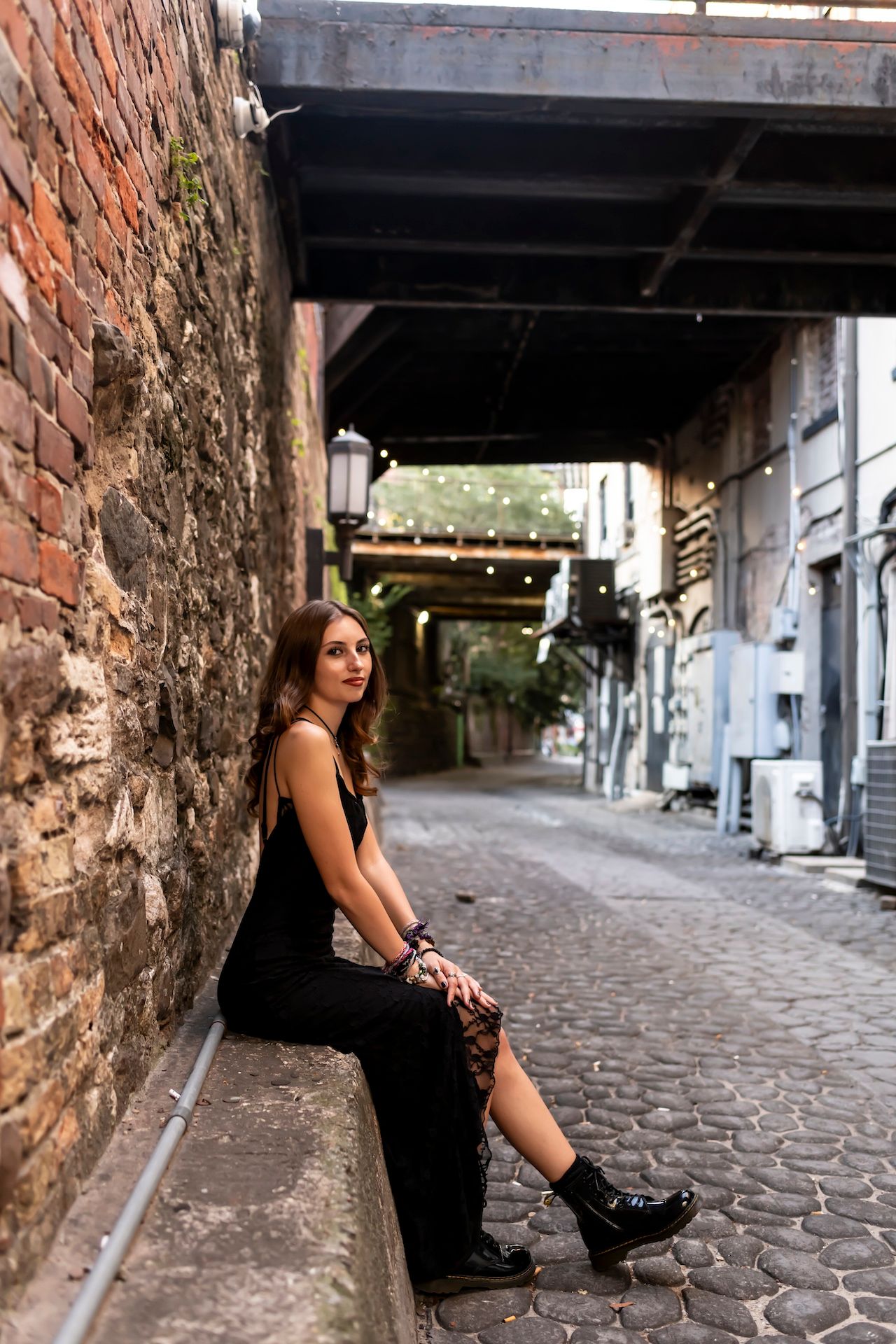 Someone in a black dress sits along a brick wall under a dark overpass on a cobblestone street in an urban setting.