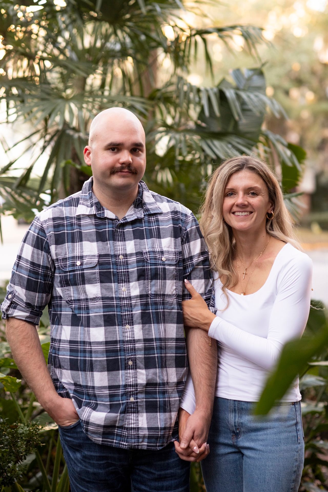 A couple in coordinating plaid shirt and white top share a romantic moment in front of tropical foliage.