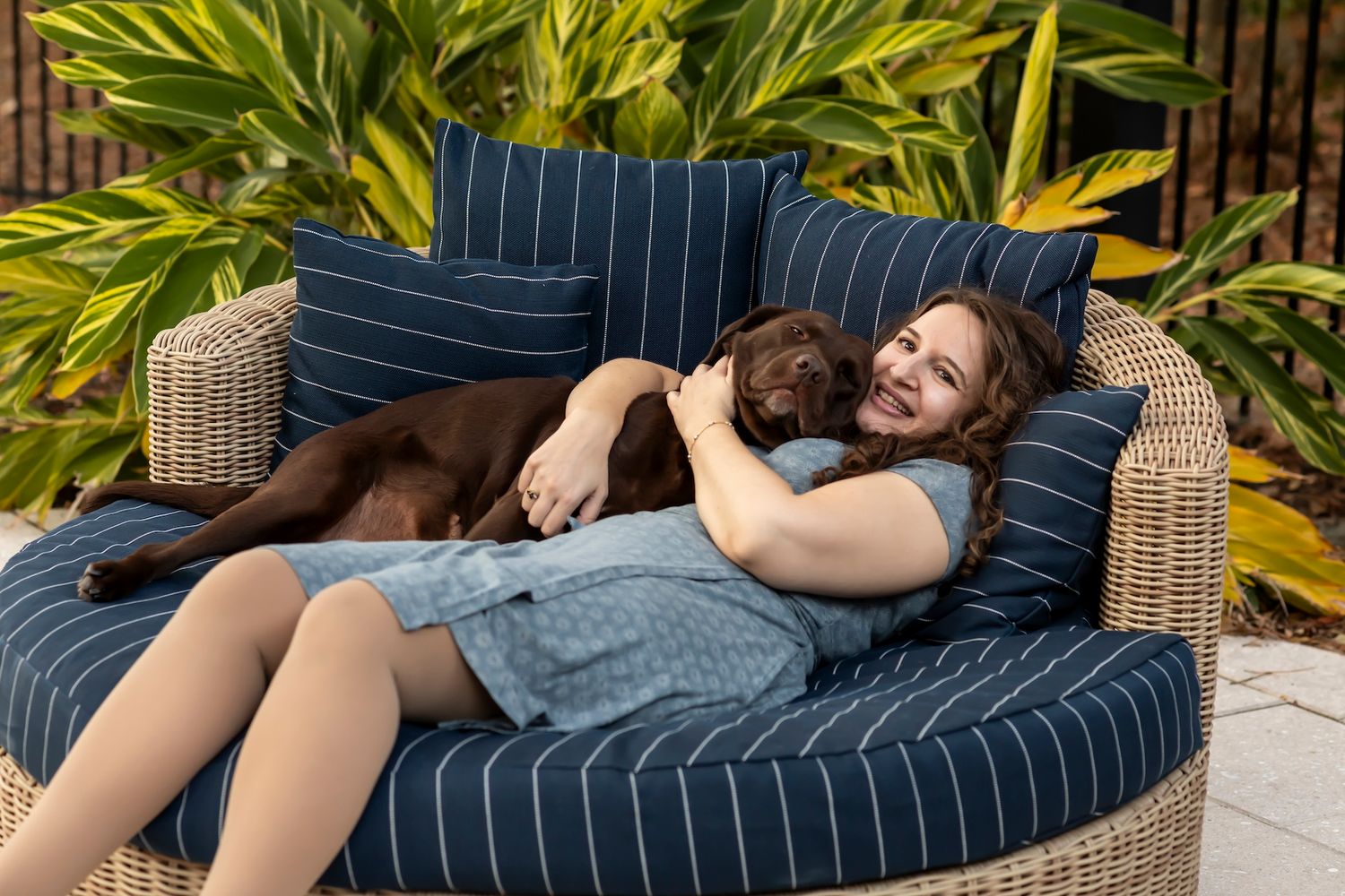 A person relaxes on a navy striped outdoor lounge chair with a dog while surrounded by tropical plants.