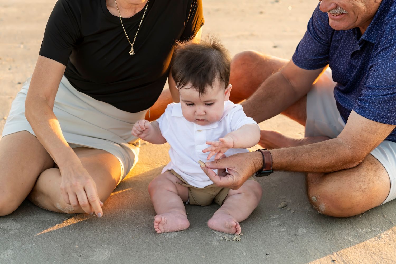 A small baby in white clothing sits on sandy beach supported by caring hands.