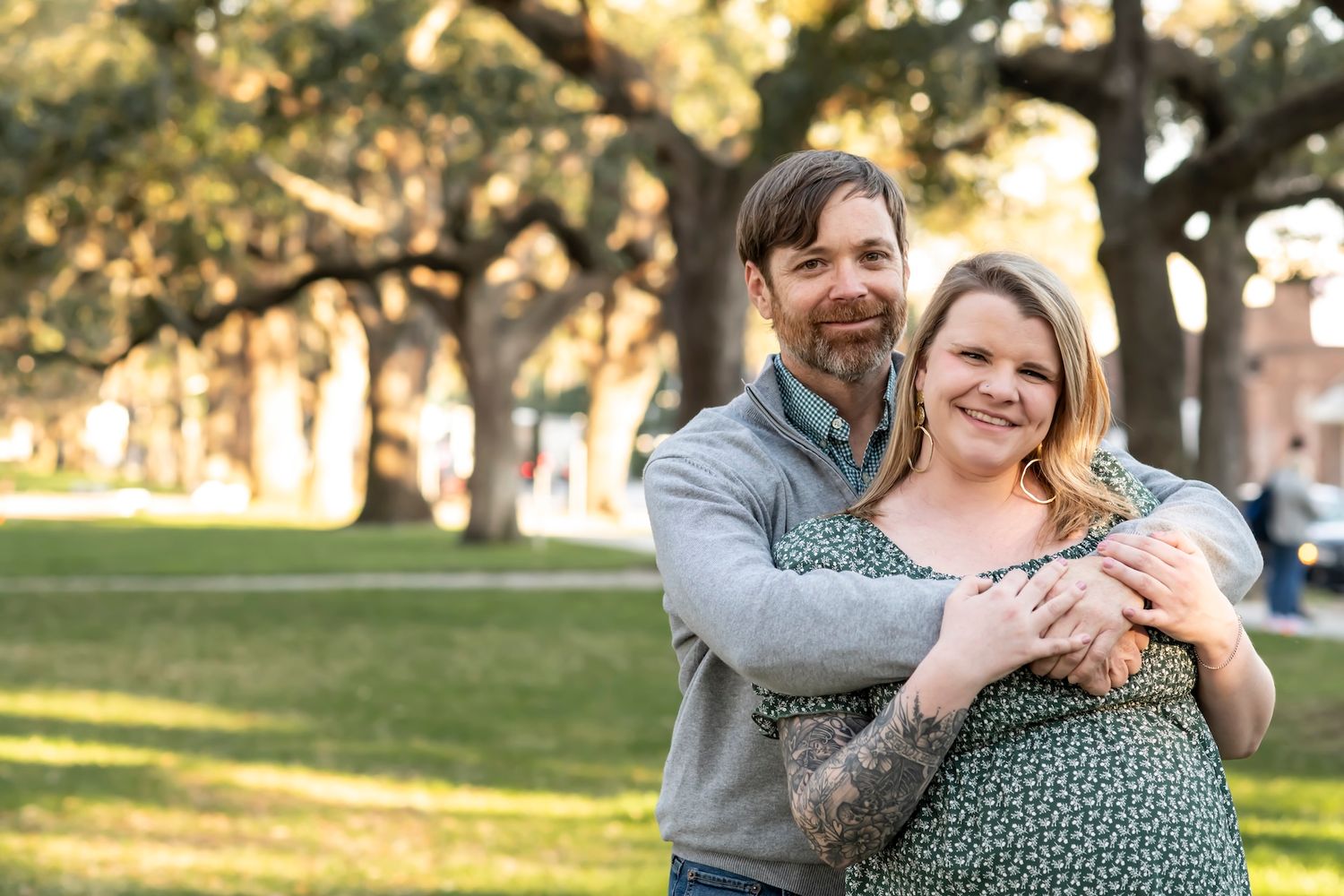A couple embraces outdoors in a romantic setting with sunlight filtering through oak trees in the background.