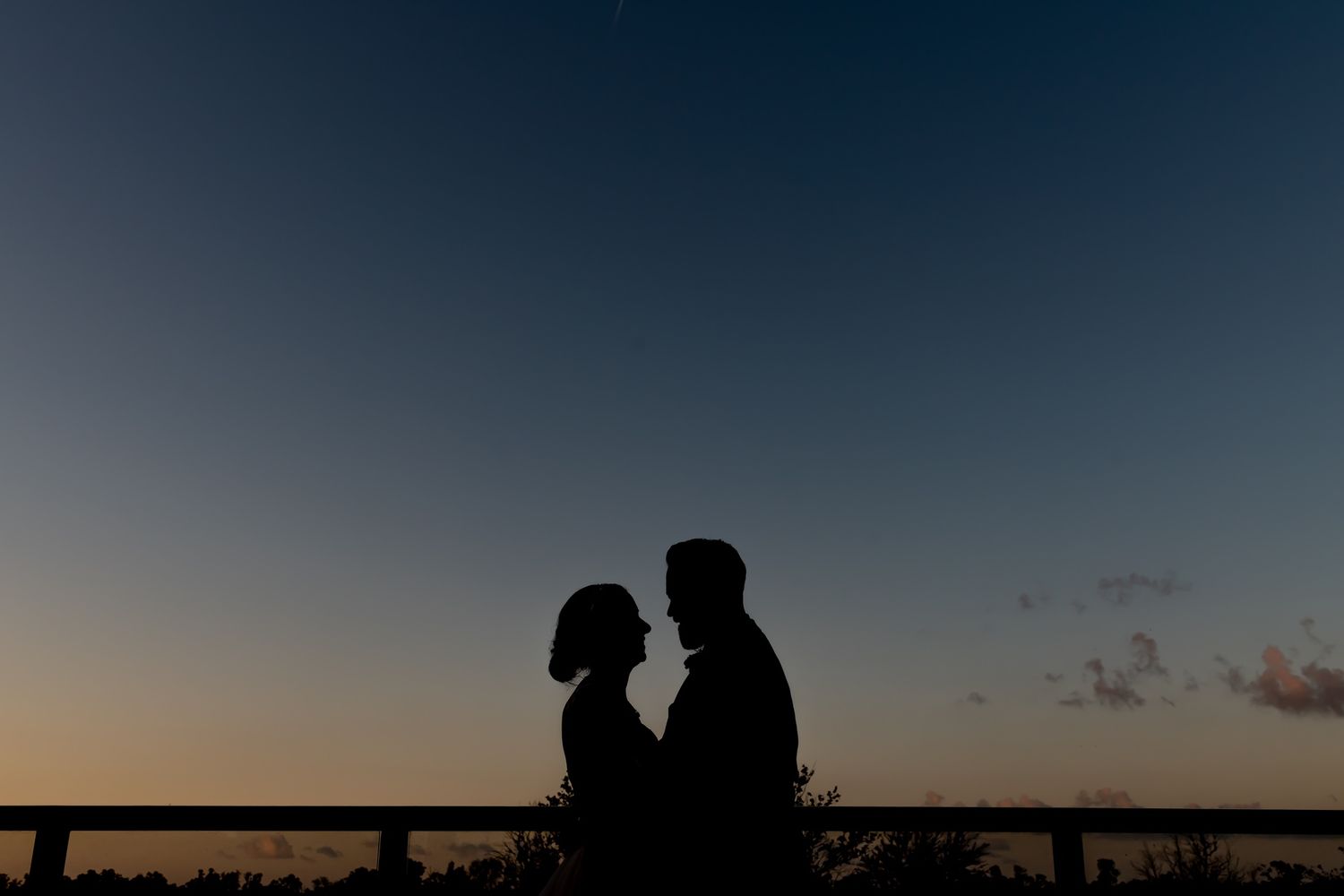 Silhouetted couple sits together on a railing against a dusky sunset sky.