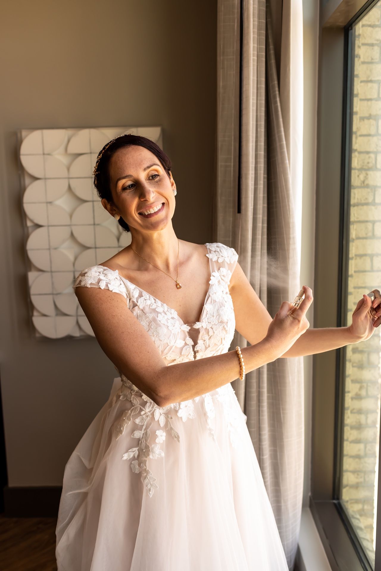 A person in a white lace wedding dress poses near a window with a cheerful expression.
