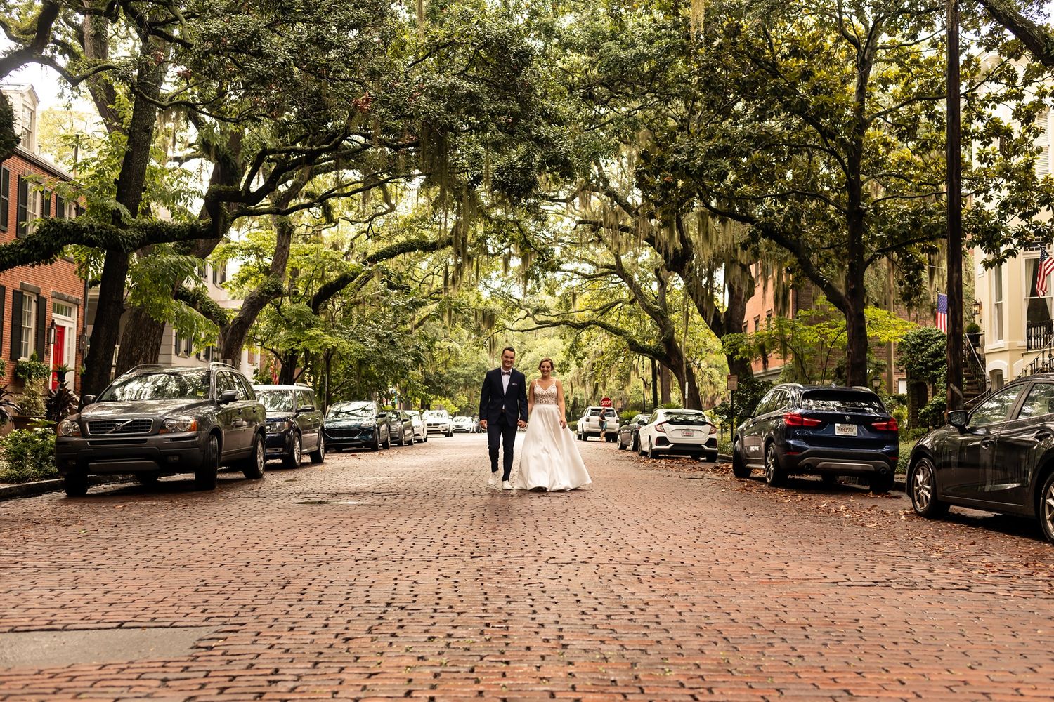 A couple walks down a picturesque cobblestone street lined with parked cars and oak trees draped in Spanish moss.