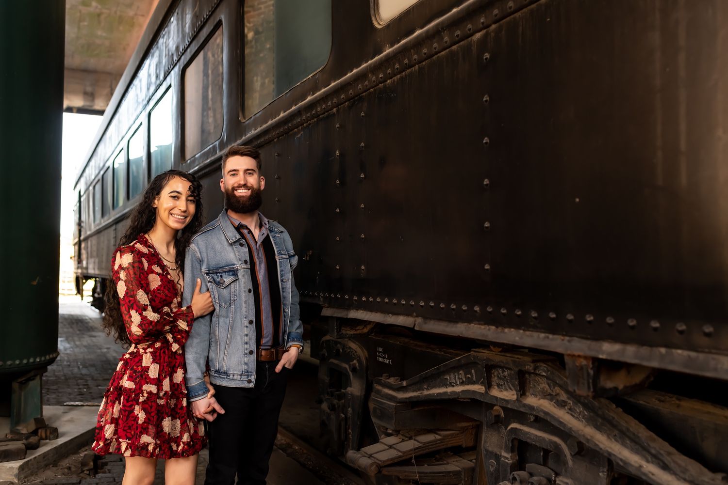 A couple in vintage-inspired outfits poses romantically at a historic train station for an engagement photo session.