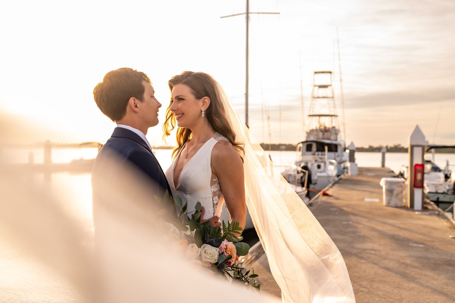 A couple shares an intimate moment along a dock during sunset with boats in the background.