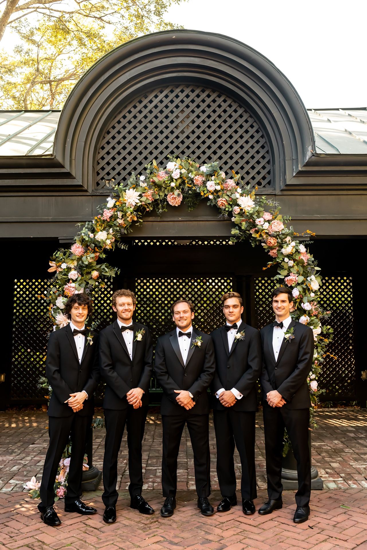 Groomsmen in black tuxedos pose under an elegant floral arch with lattice background at an outdoor wedding venue.