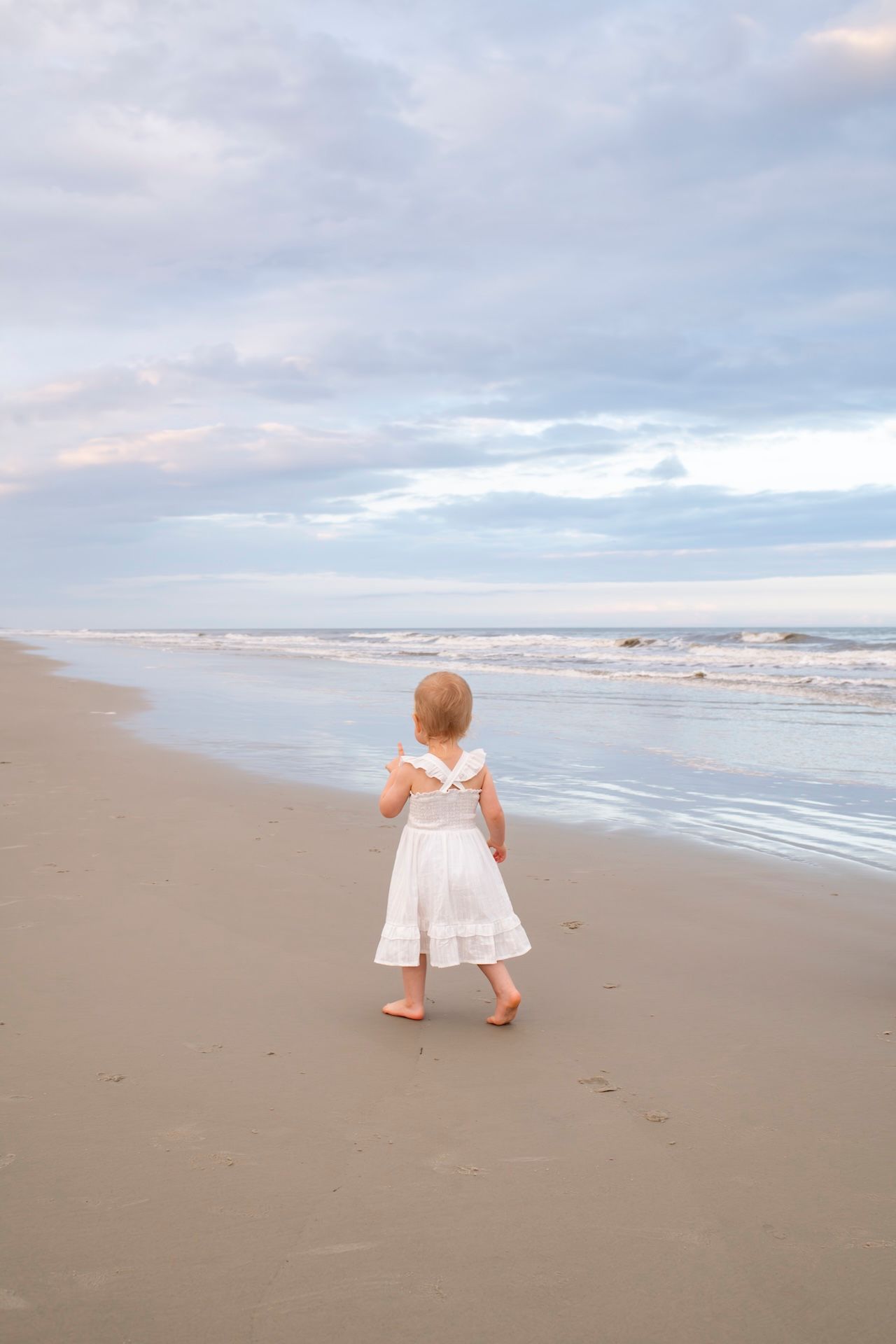A young girl in a white dress walks barefoot along a sandy beach toward gentle ocean waves under a cloudy sky.