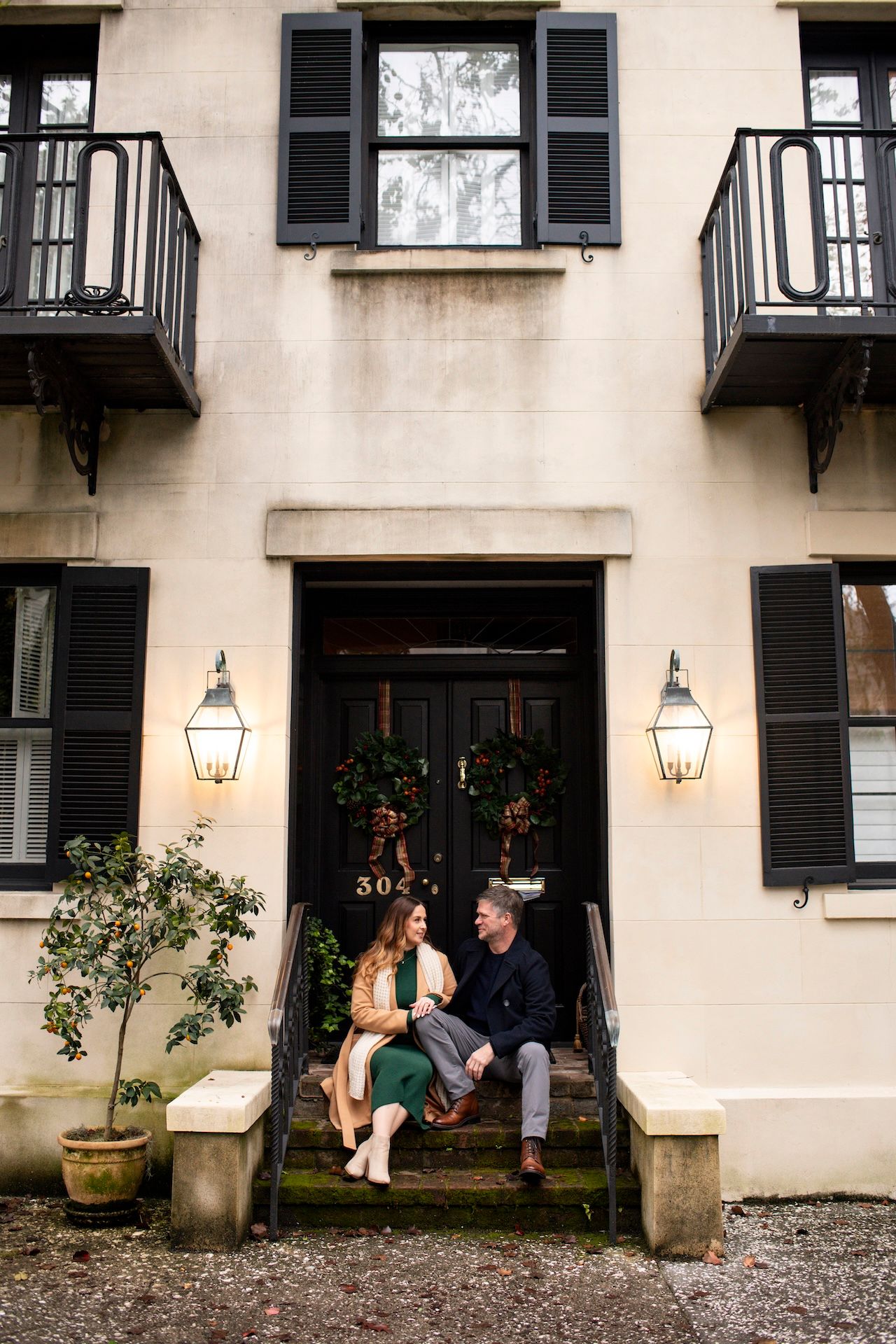 Couple sitting on steps of elegant cream building with black shutters, wreaths, and lanterns at dusk.
