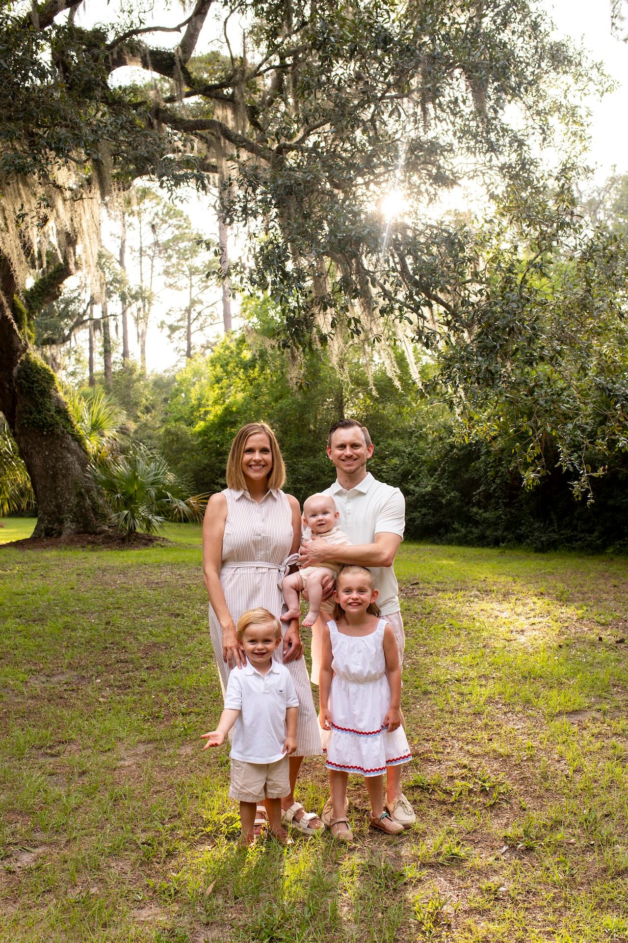 Family of five posing outdoors under a large oak tree with Spanish moss in a lush green park setting.
