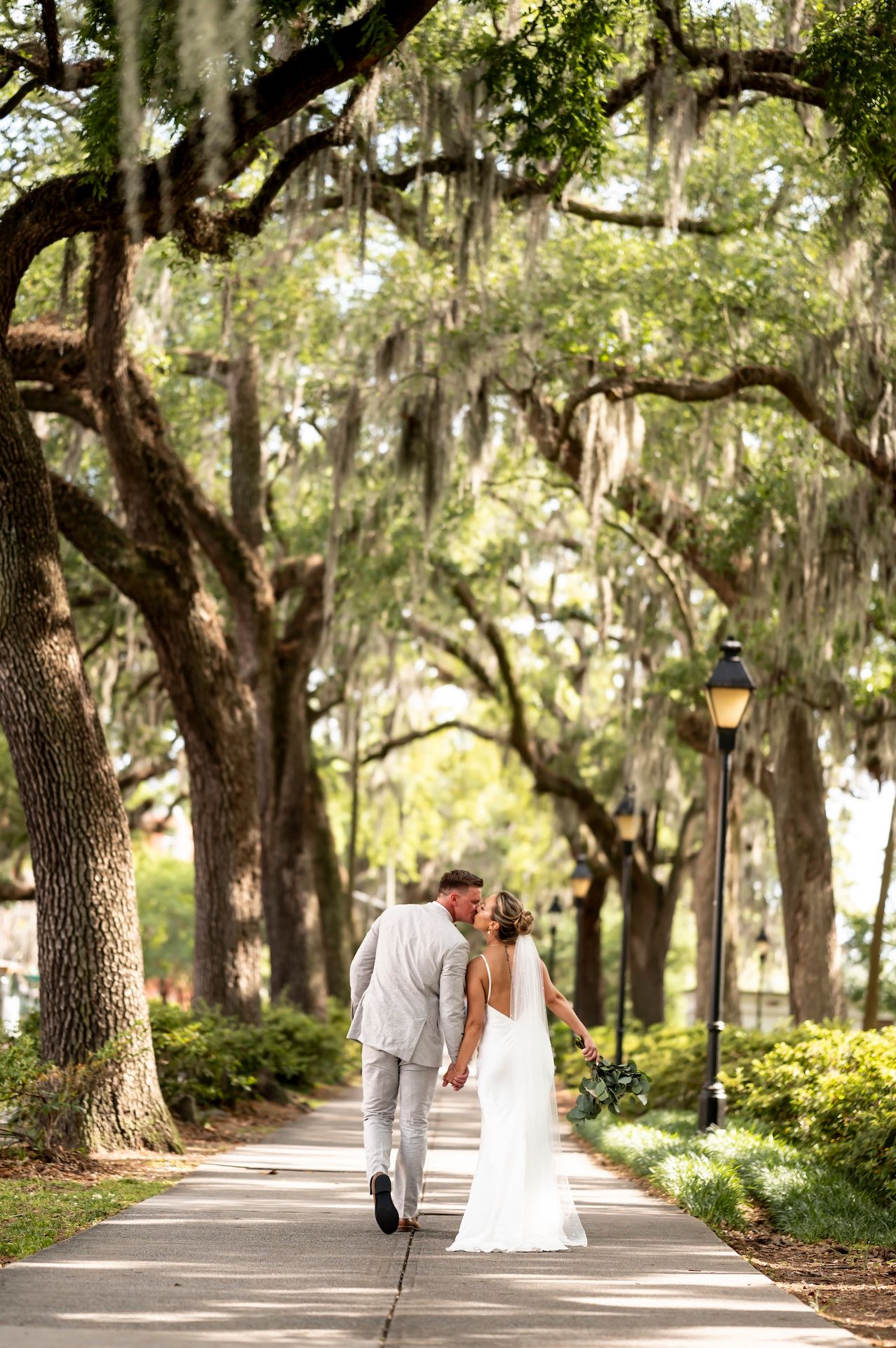 A tree-lined pathway with Spanish moss hanging from ancient oaks with a bride and groom kissing at Forsyth Park.
