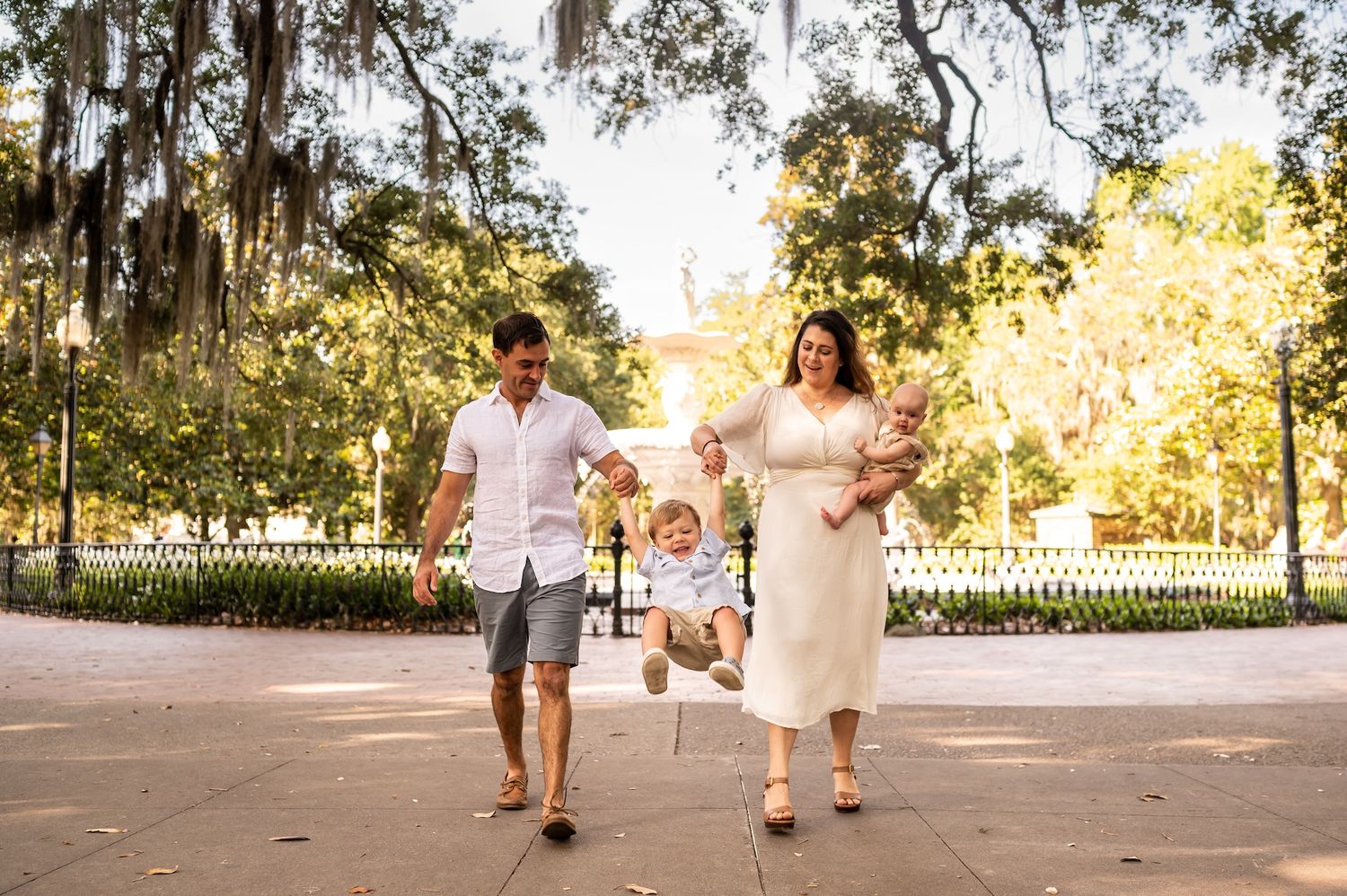 A family walks together in Forsyth park in front of the fountain with Spanish moss hanging from trees overhead.
