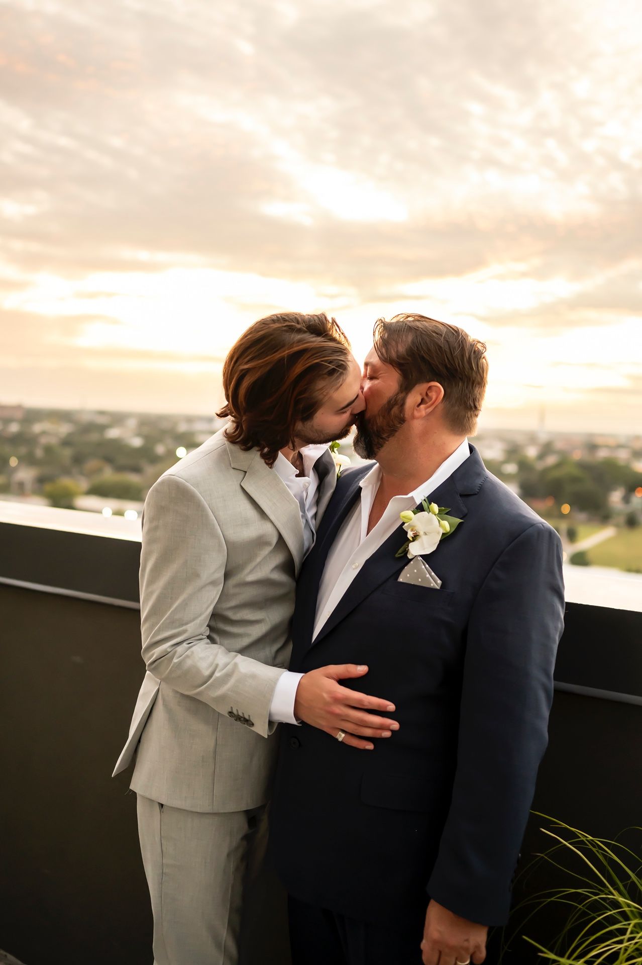 Two people in wedding attire share an intimate moment during sunset on a rooftop overlooking a city landscape.