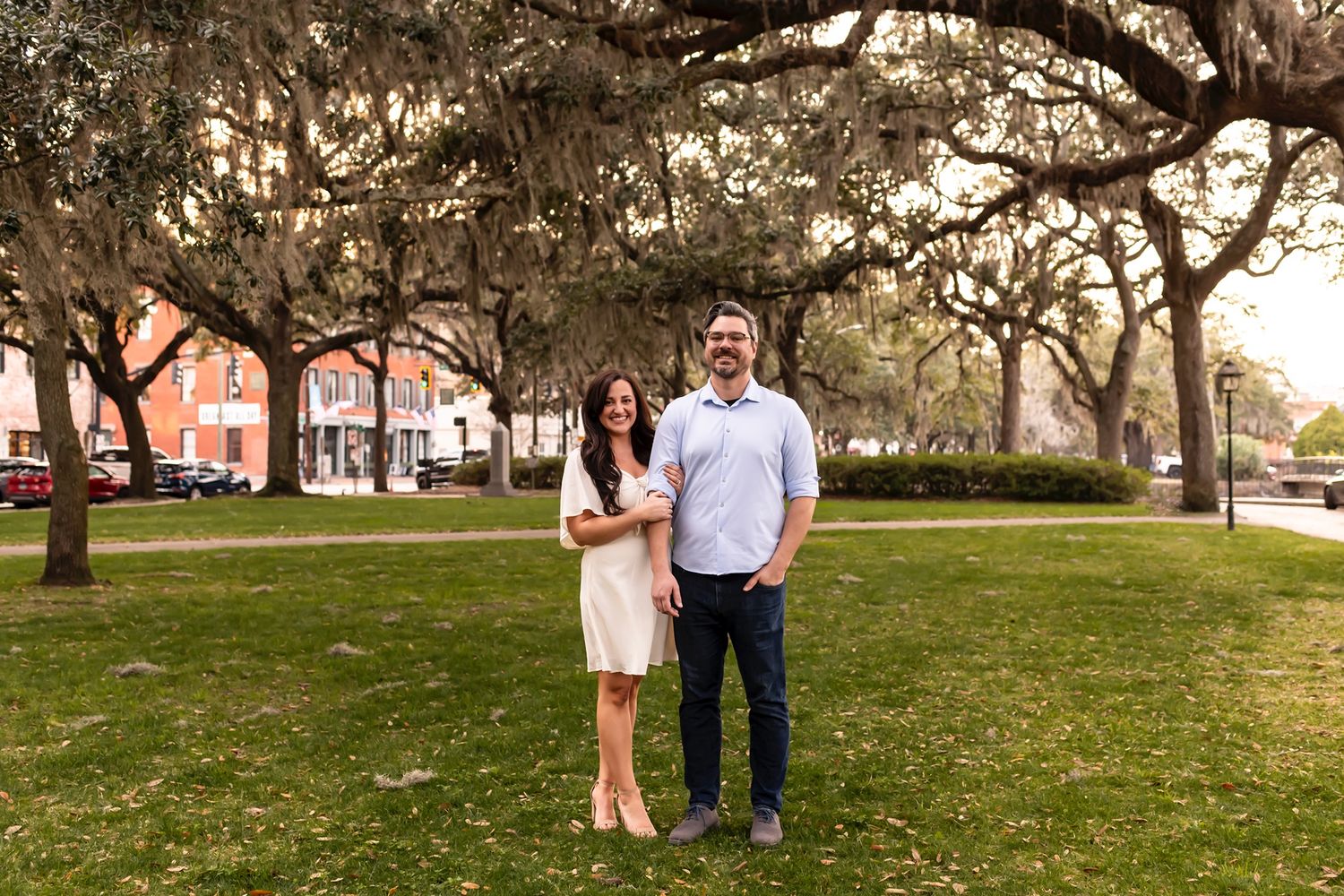 A couple stands together on grass beneath Spanish moss-draped oak trees in a scenic park at sunset.