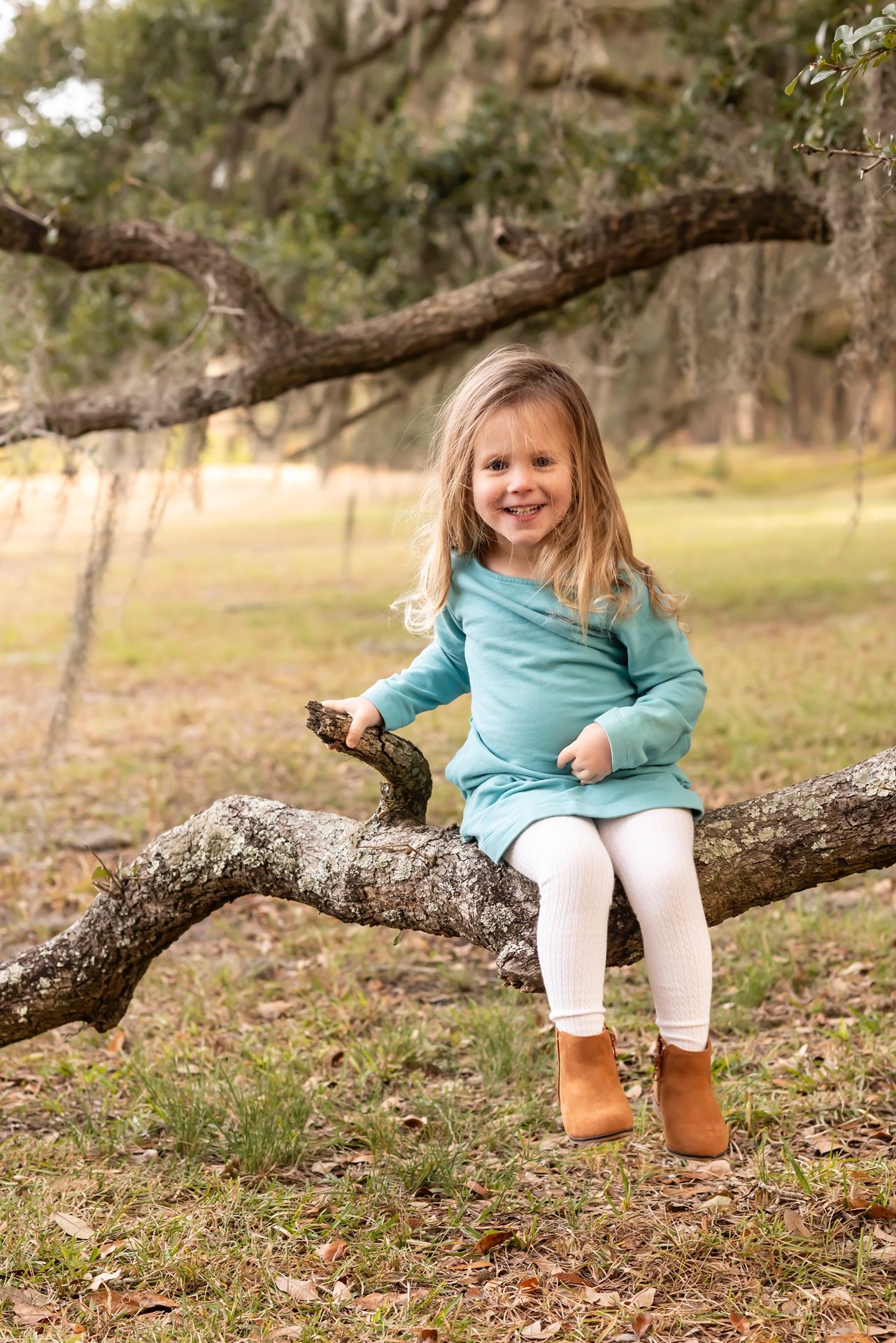 A child wearing a turquoise sweater and white leggings sits on a fallen tree branch on an autumn day.